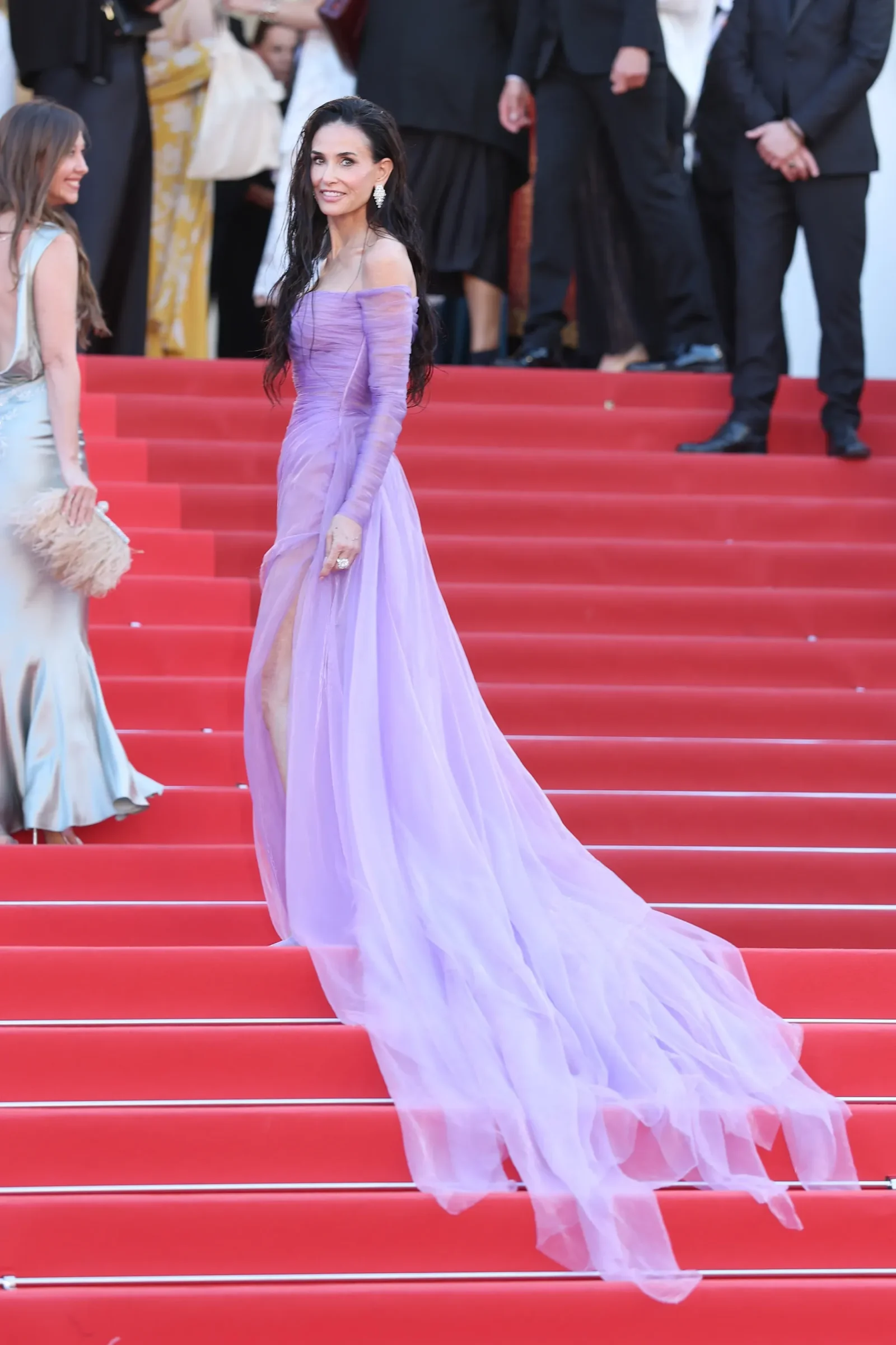 Elegant woman in an off-shoulder lavender gown descending red-carpet stairs at a formal event, herself posing for photos with crowd behind.