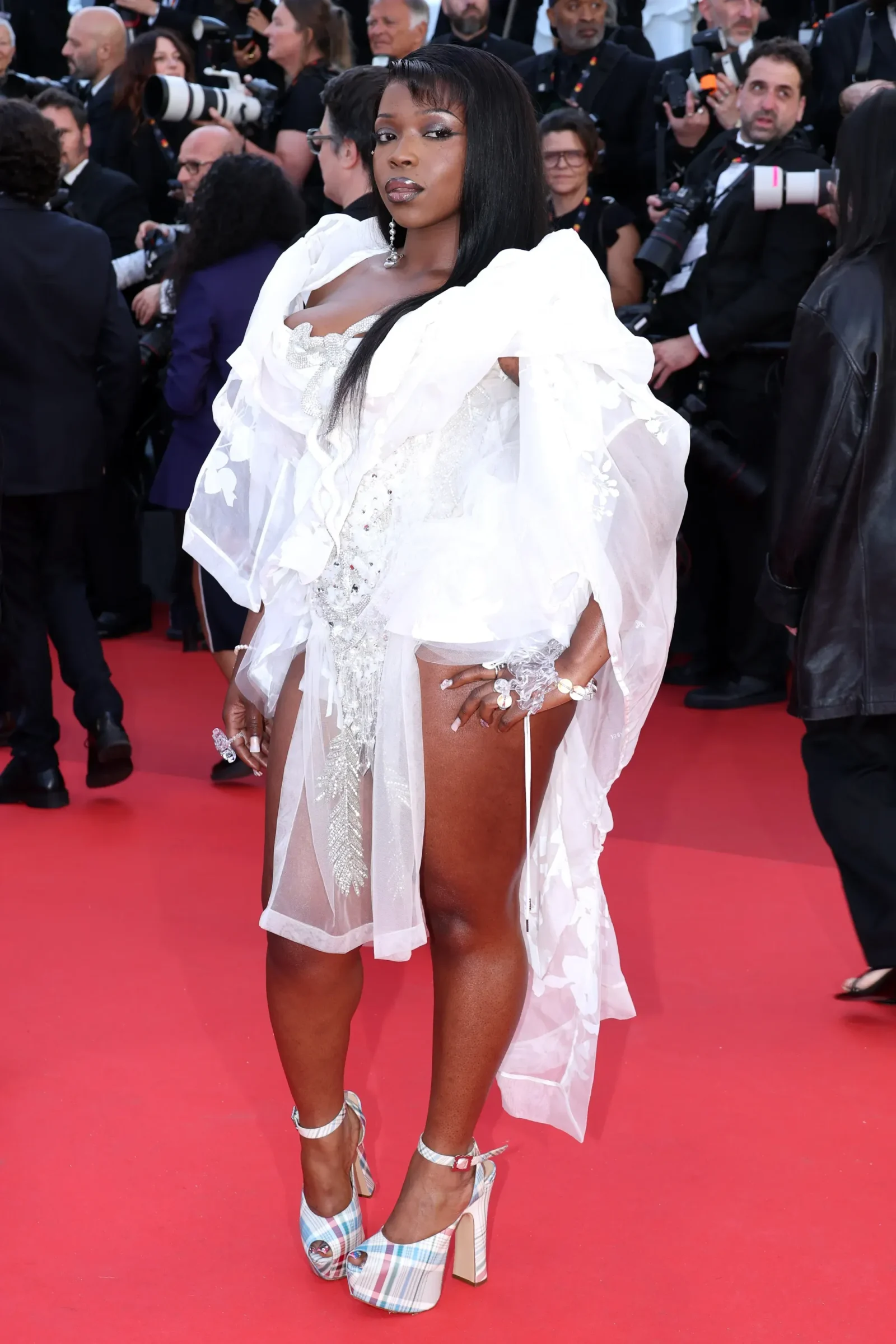 Woman on a red carpet wearing a white ruffled gown with silver jewelry and plaid platform heels, photographers in the background