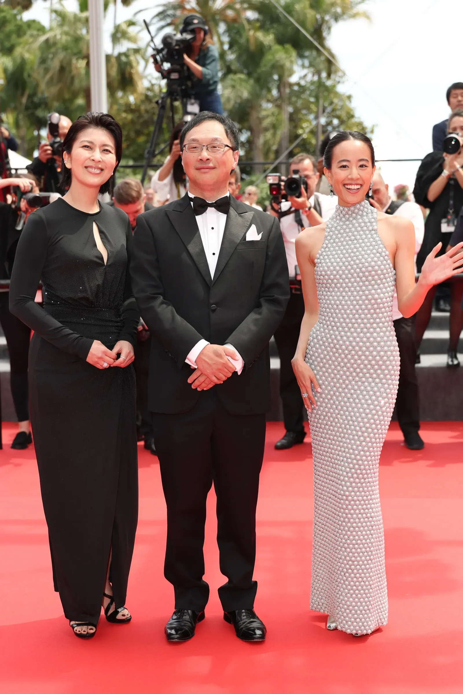 Three celebrities pose on a red carpet: a woman in a black gown on the left, a man in a black tuxedo center, and a woman in a silver beaded gown on the right, with photographers in the background.