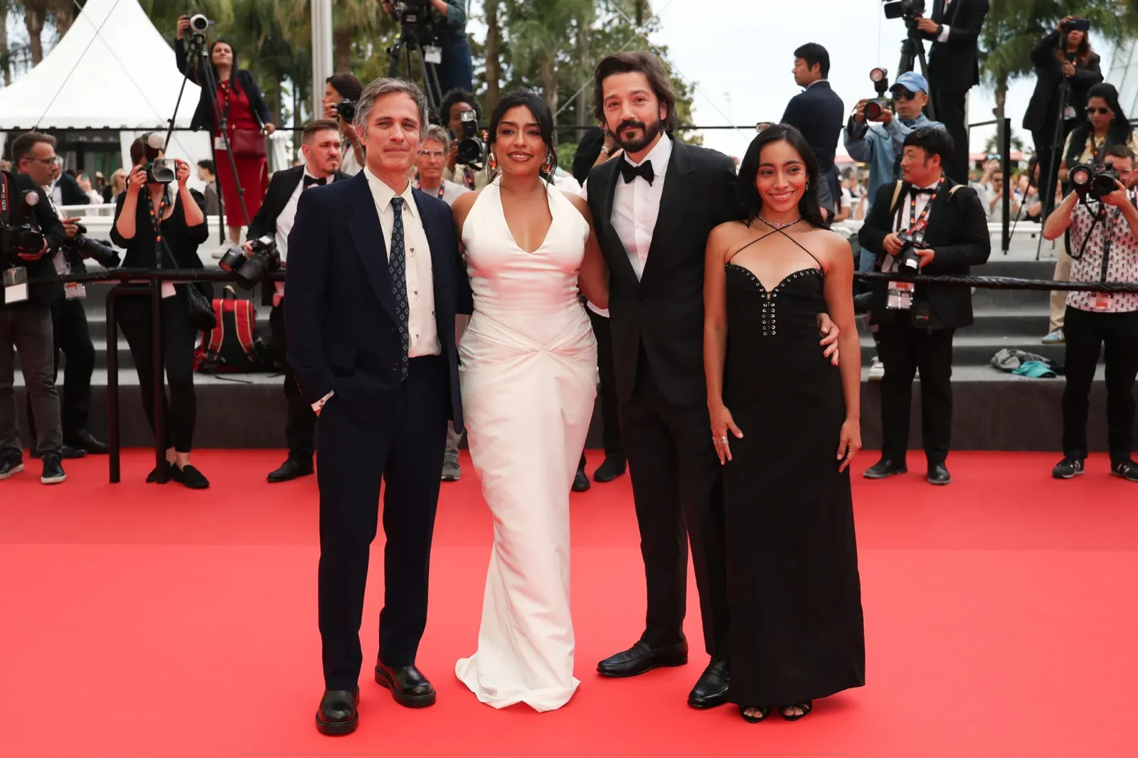 Four people posing on a red carpet as photographers surround them, smiling for the cameras. The woman second from left wears a white gown, flanked by a man in a navy suit and another man in a black tux, with a woman in a black dress on the far right.