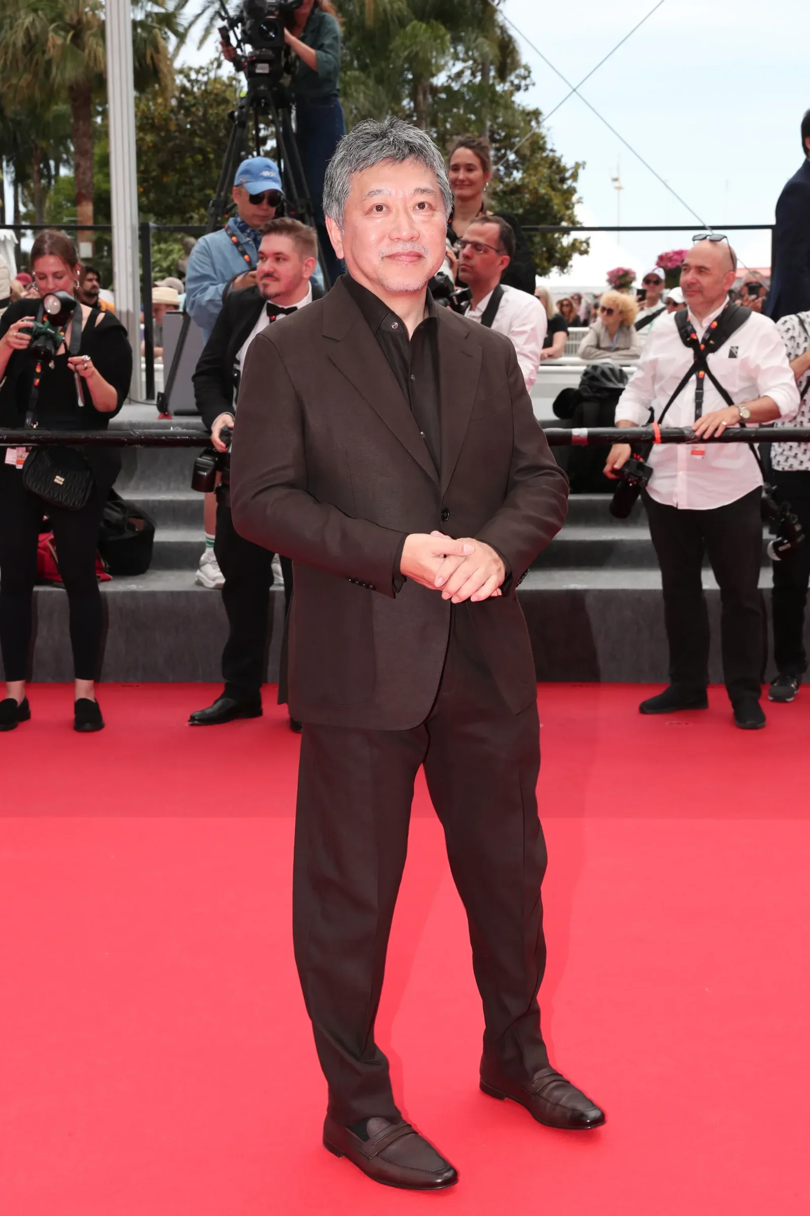 Man in a dark suit stands on a red carpet facing the camera, with photographers and crew in the background at a media event outdoors.