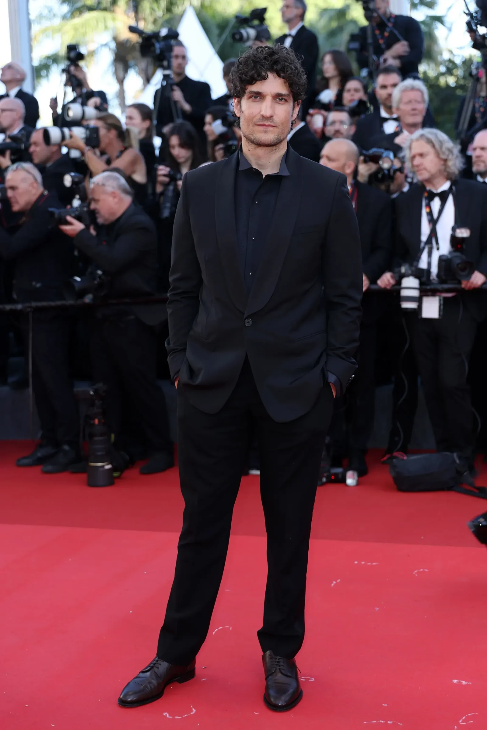 Man in a black suit standing on a red carpet as photographers crowd the background, holding cameras and taking photos.