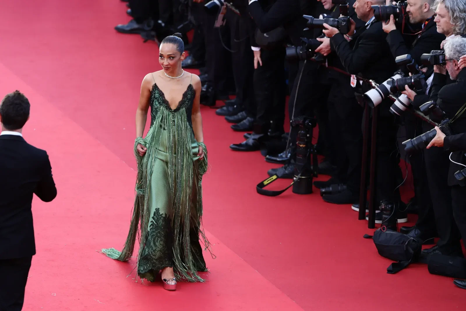 Woman on a red carpet wearing an emerald-green fringe gown, posing for photographers on a line of cameras to the right.