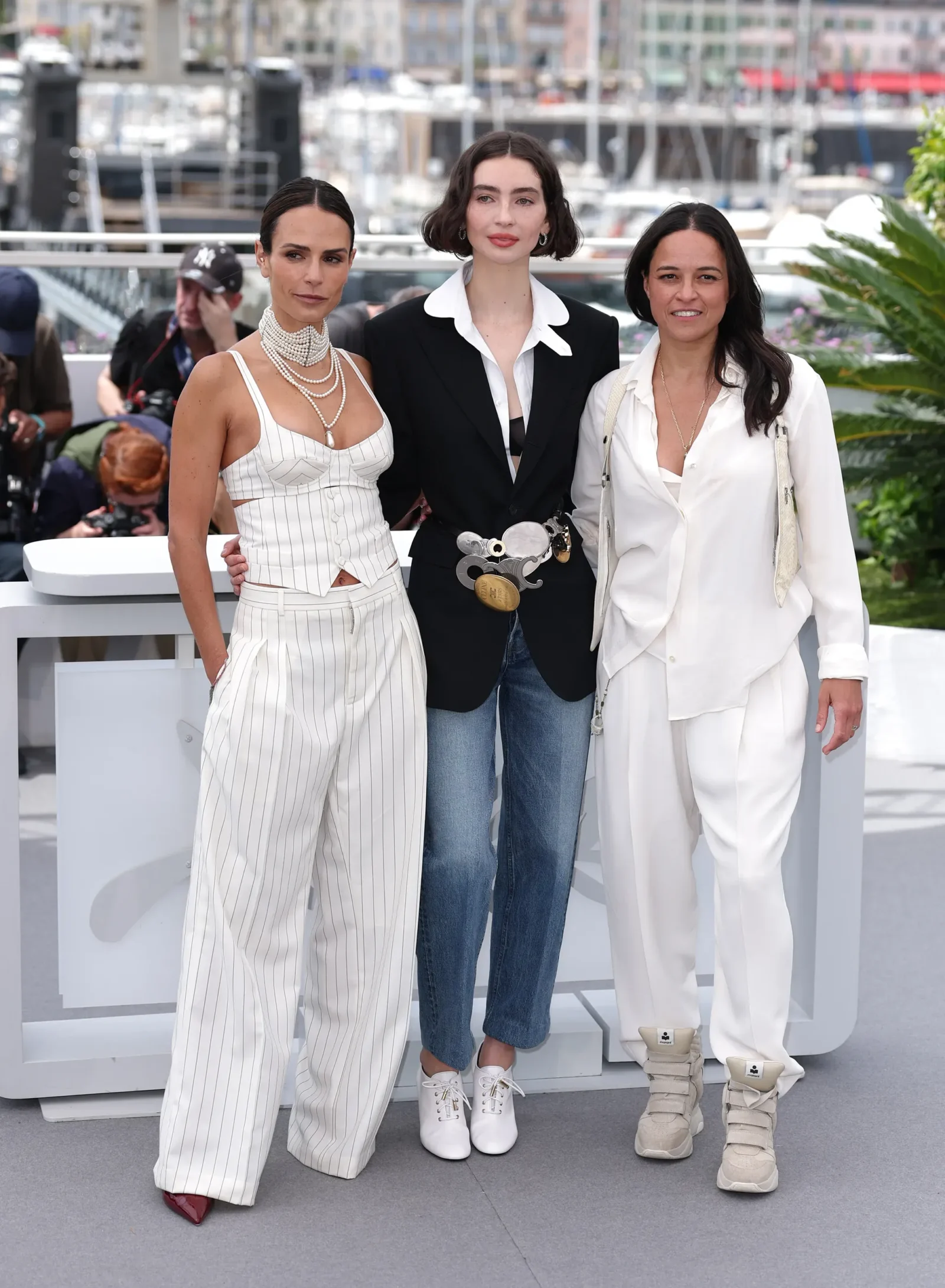 Three fashionable women posing together at a marina event; left wears a white pinstripe suit with layered pearl necklaces, middle in a black blazer with a bold metallic belt, and right in an all-white outfit with oversized boots and a cream bag, outdoors by boats and greenery
