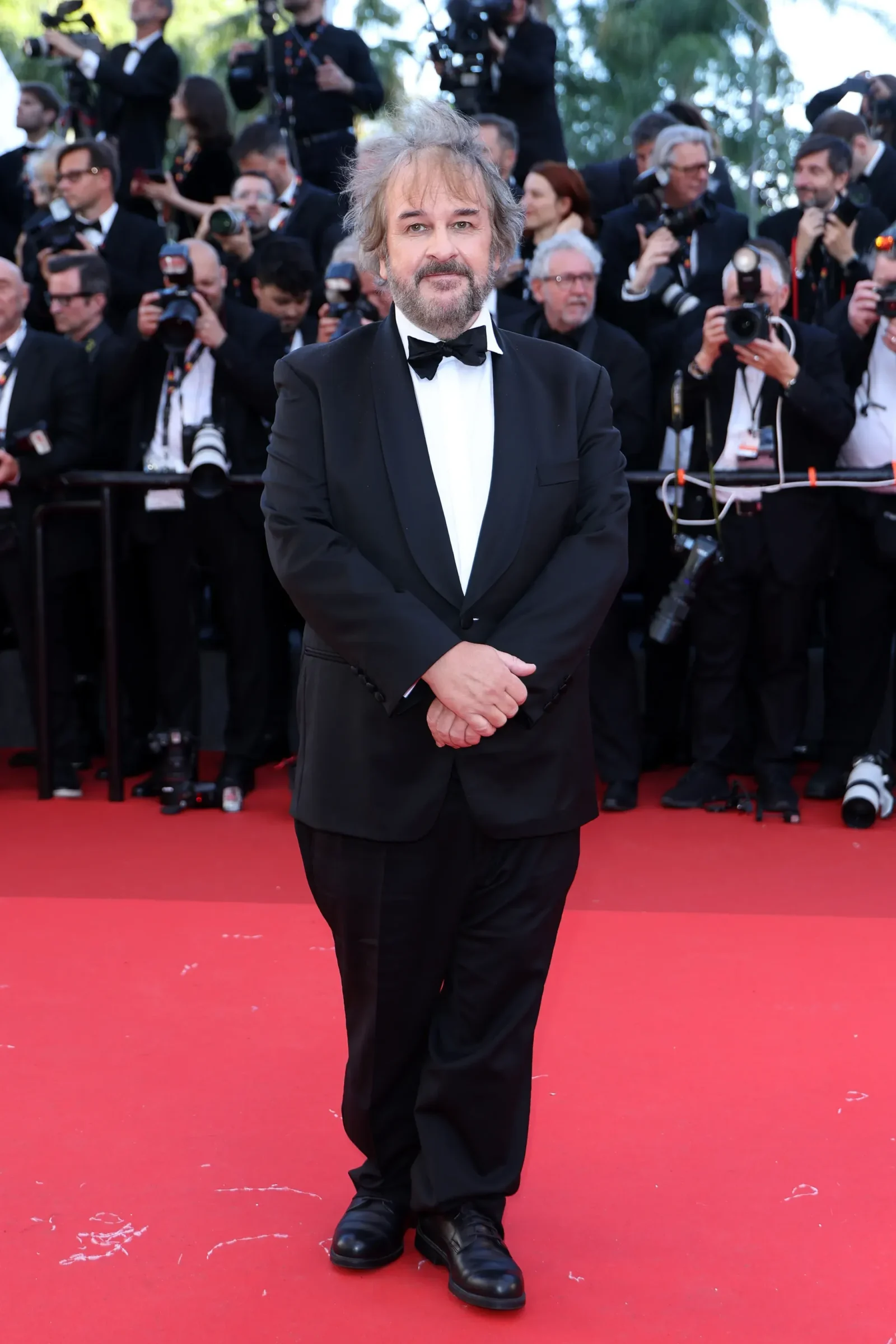 Man in a black tuxedo posing on the red carpet as paparazzi crowd him with cameras behind.