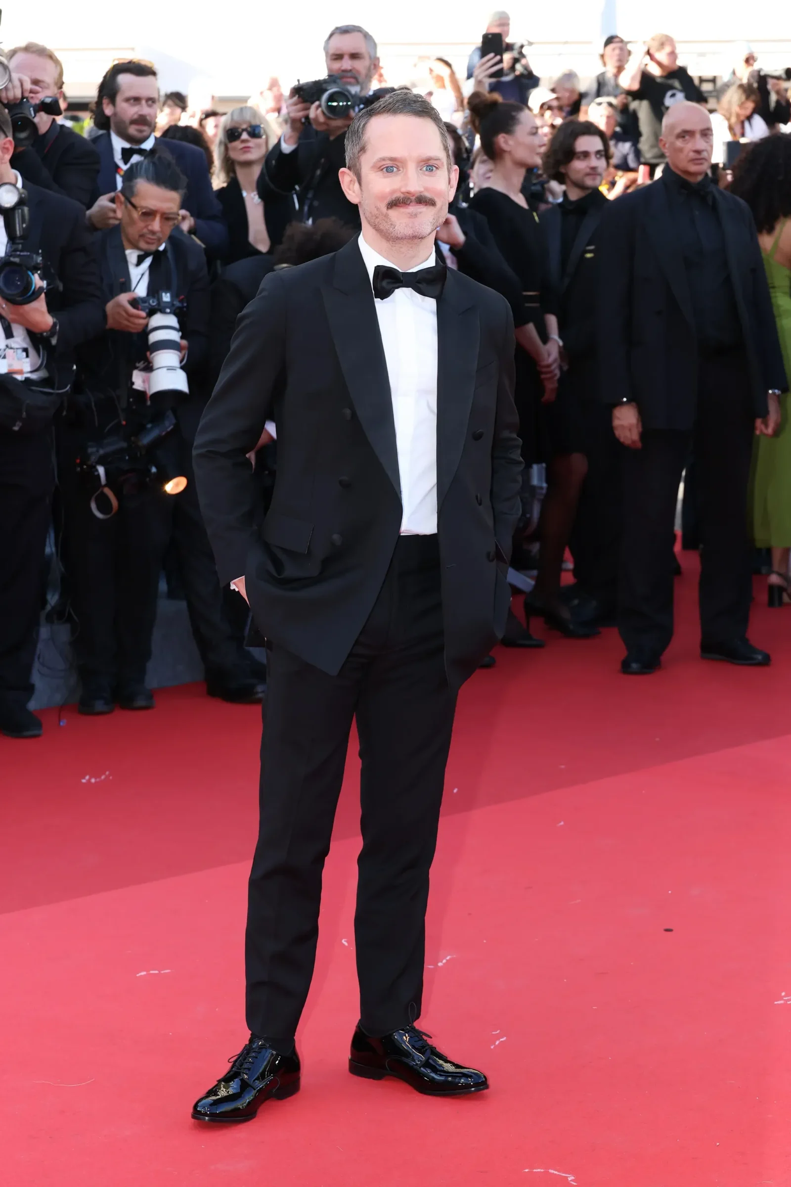 Man in a black tuxedo and bow tie posing on a red carpet with photographers and onlookers behind him.