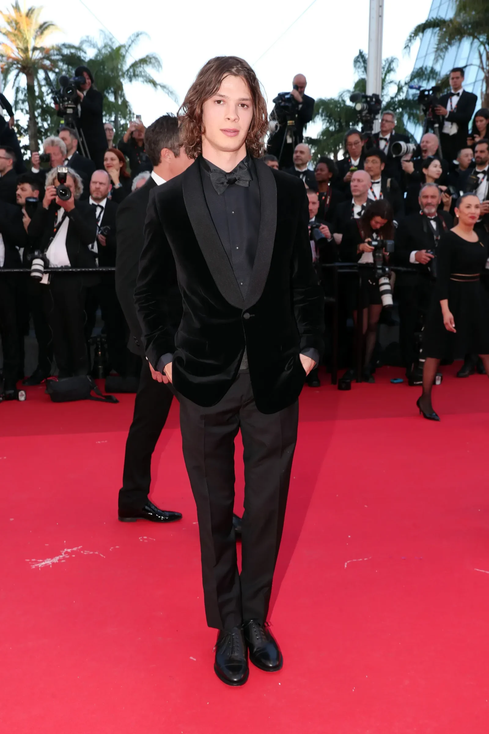 Young man in a black velvet tuxedo with bow tie on a red carpet, photographers in the background capturing the moment.