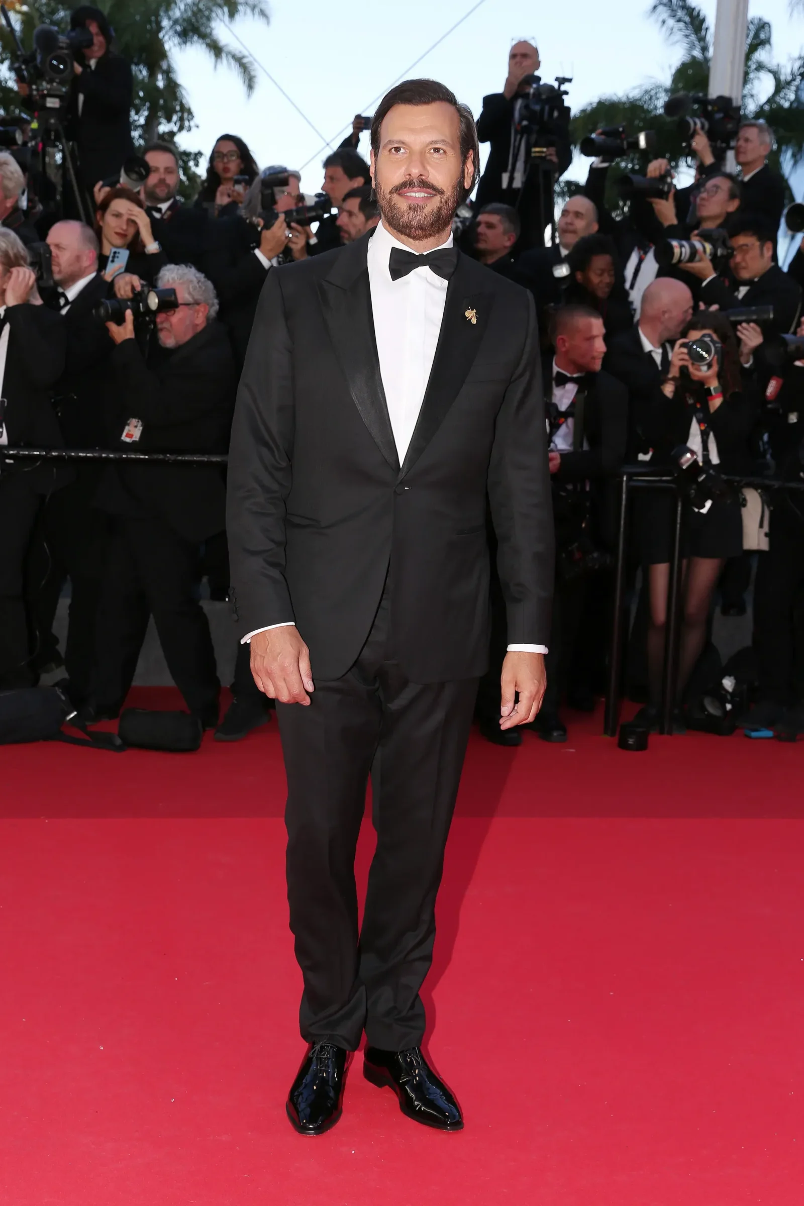 Man in a black tuxedo posing on a red carpet with photographers in the background