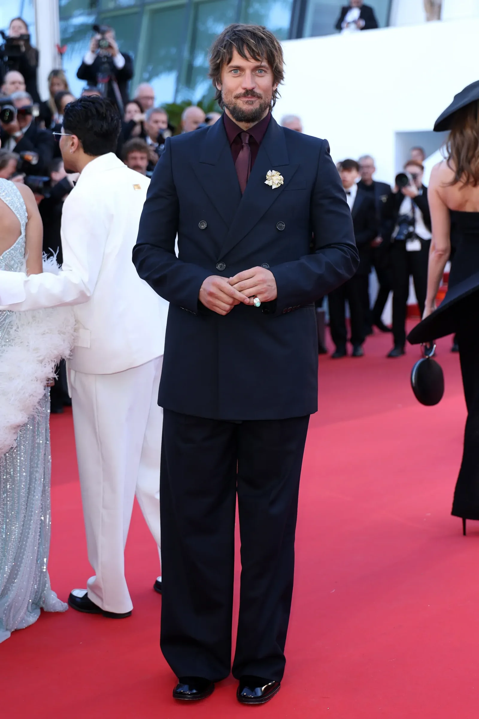 Man in a double-breasted navy suit with a maroon shirt and tie, standing on a red carpet with photographers in the background; lapel boutonnière visible on left breast.
