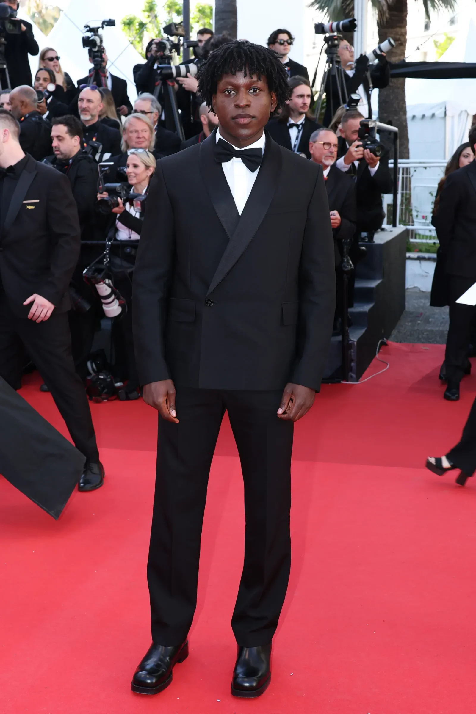 Young man in a black tuxedo and bow tie standing on a red carpet, with photographers in the background at a formal event.