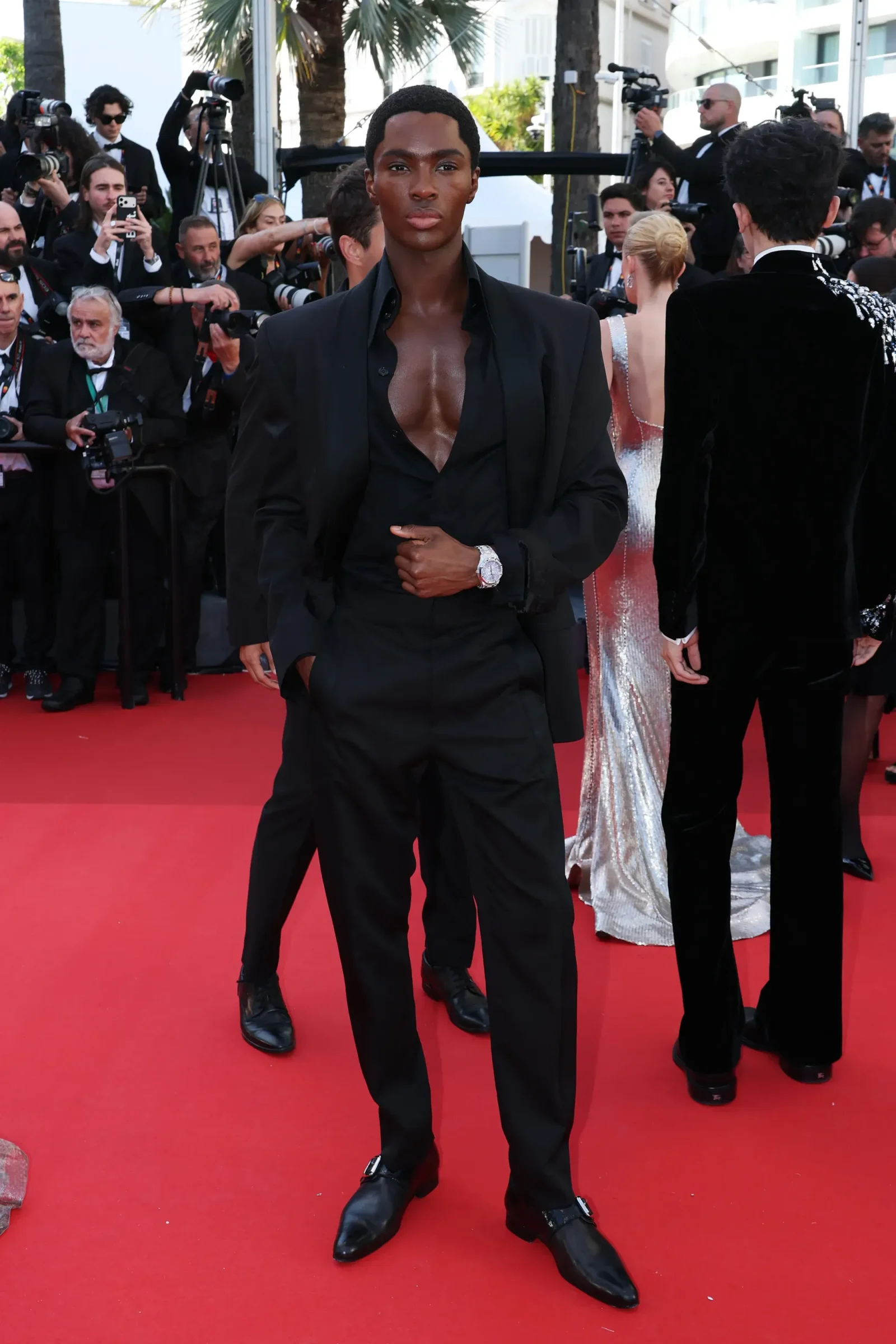 Man in an open-black suit and unbuttoned shirt on a red carpet, surrounded by photographers at a formal event.
