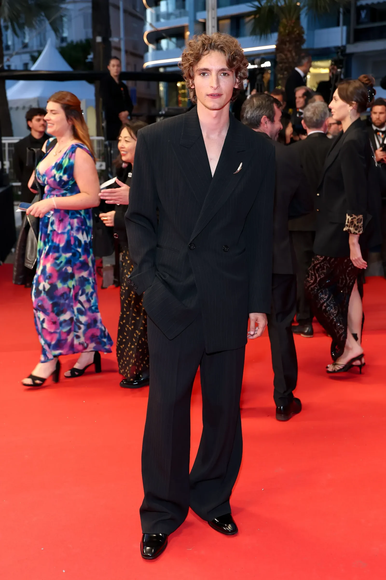 Man on a red carpet wearing a dark pinstripe double-breasted suit with hands in his pockets, guests in formal attire in the background.