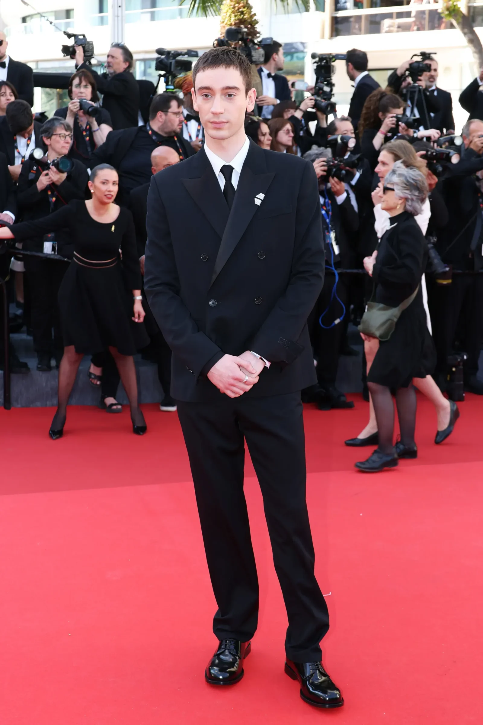 Young man in a black double-breasted suit and tie posing on a red carpet at a media event, surrounded by photographers.