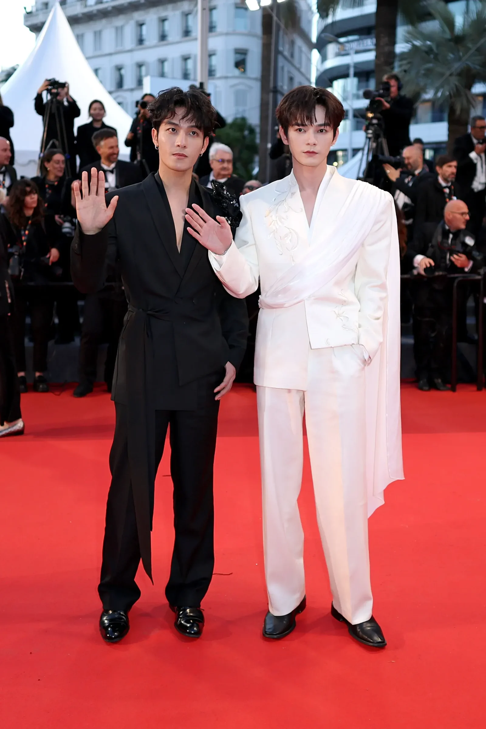 Two men posing on a red carpet; one in a black suit waves, the other in a white embroidered suit stands beside him.