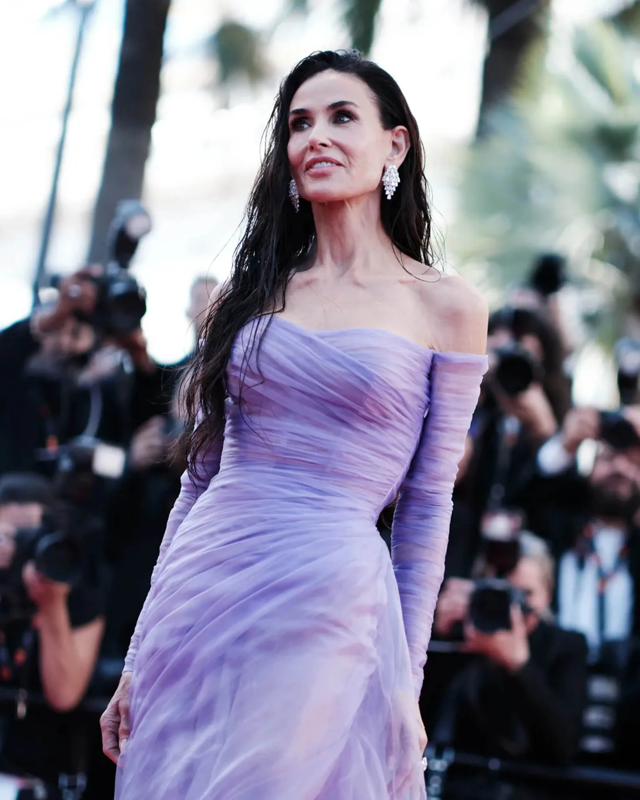 Woman in a lavender off-the-shoulder gown posing on a red carpet with photographers in the background, wearing large chandelier earrings.