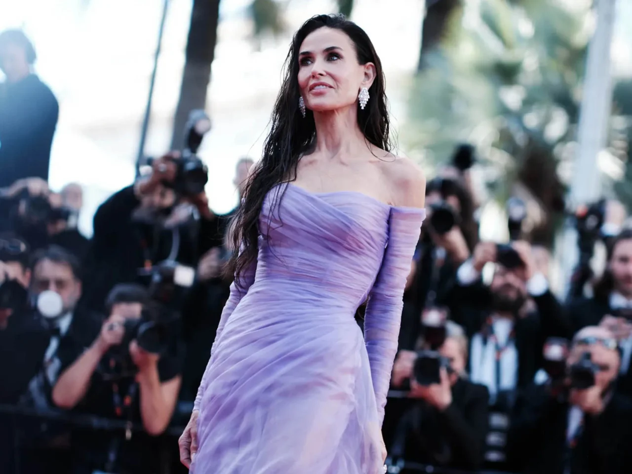 Woman in a lavender off-shoulder gown posing on a red carpet as photographers surround her.