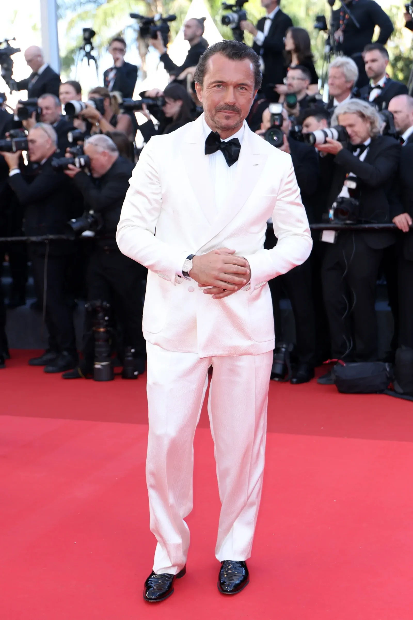 Actor in a white tuxedo posing on a red carpet as photographers surround him.