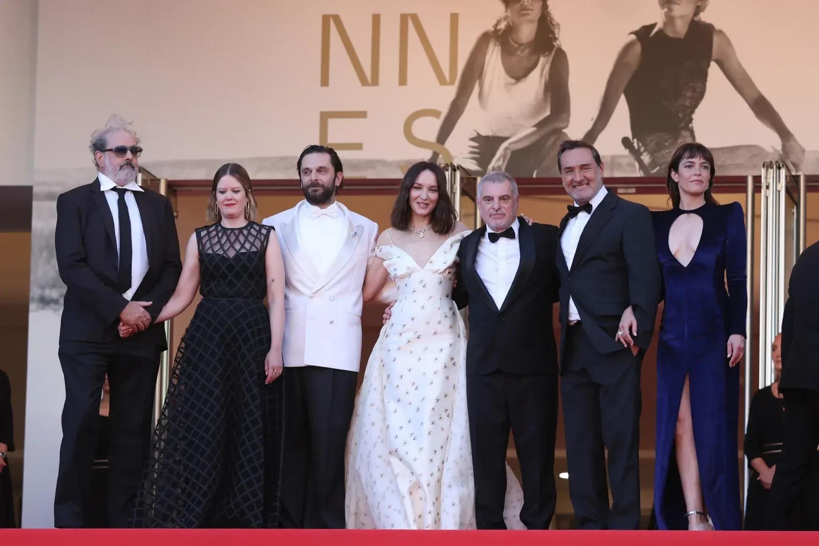 Seven formally dressed people posing on a red carpet in front of a large backdrop with fashion imagery, smiling for the camera.