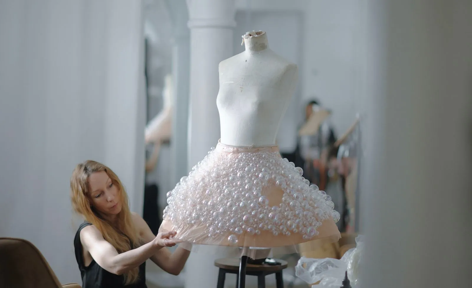 Designer adjusting a pale pink bubble-hem dress on a white mannequin in a bright studio.