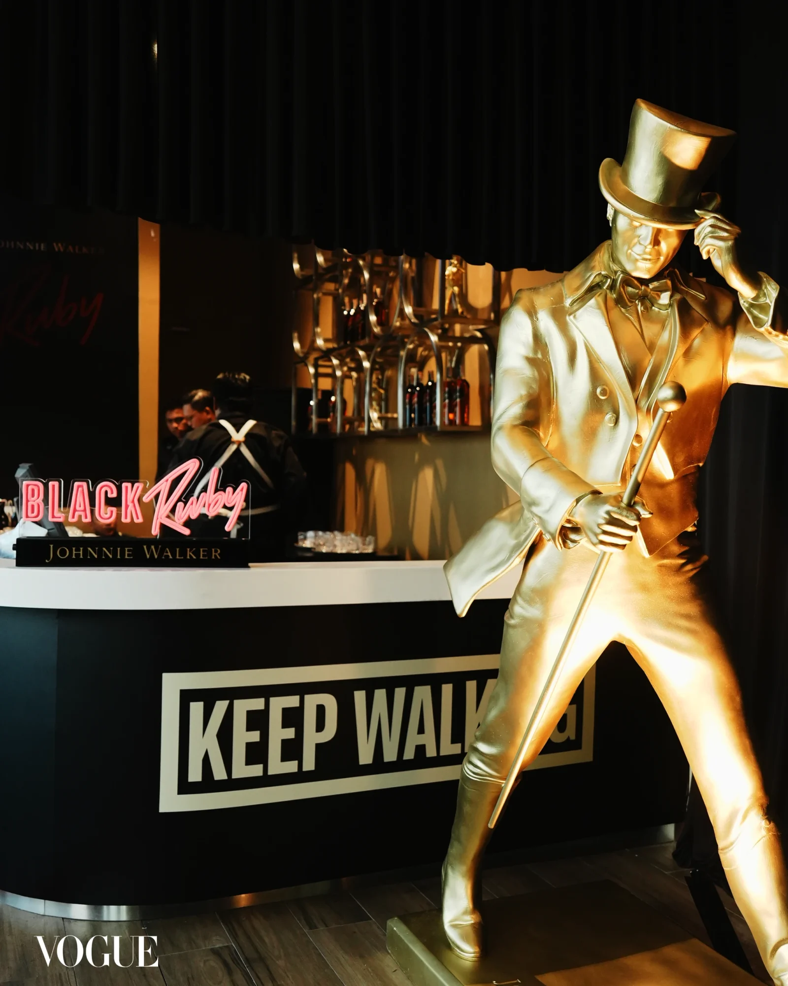 Gold statue of a man in a tuxedo and top hat, holding a cane, in a Johnnie Walker display with a bold 'KEEP WALKING' sign in the background.