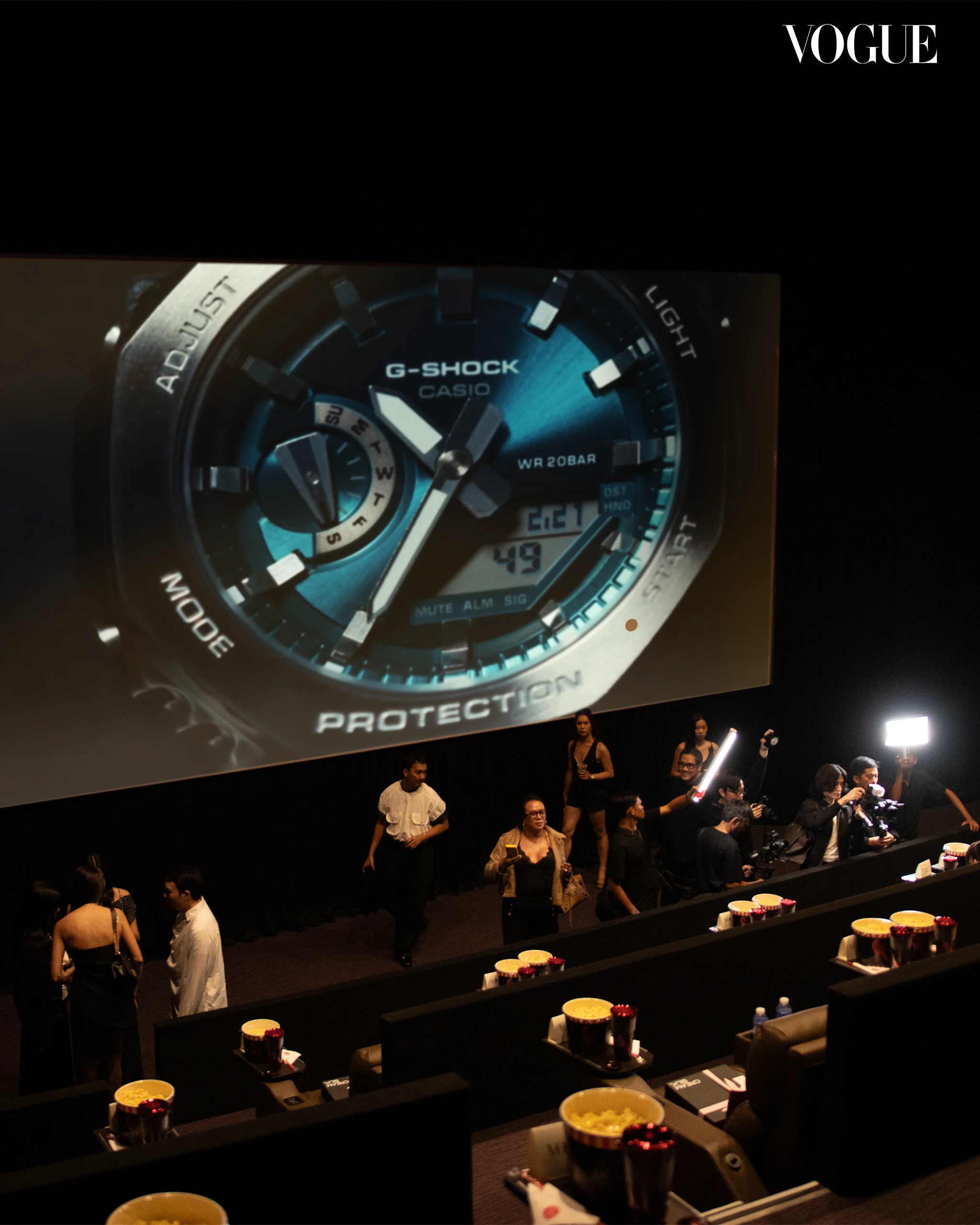 Large screen showing a blue Casio G-Shock watch at a Vogue event, with people mingling on a dark stage in the foreground.