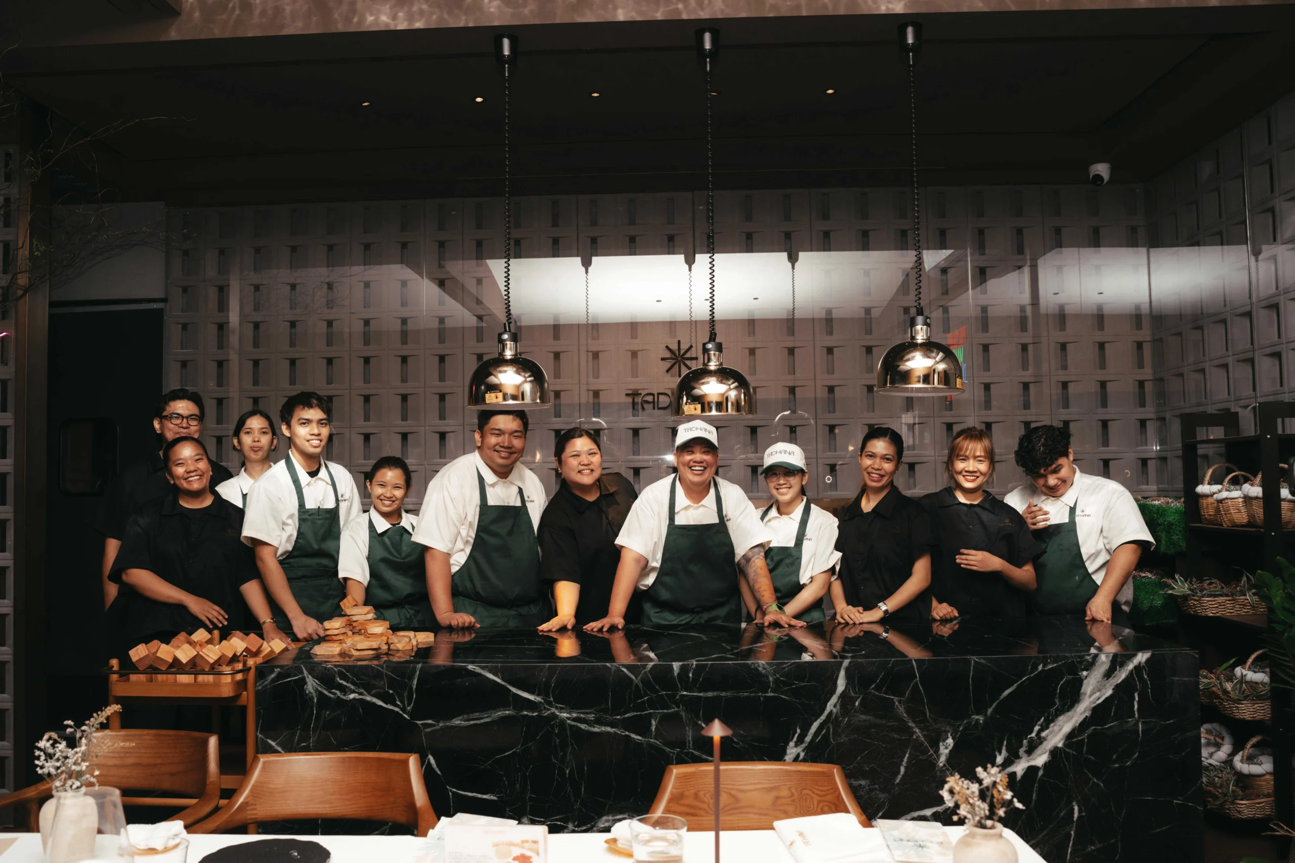 Group of kitchen staff and chefs posing behind a black marble counter with wooden bread blocks in front and pendant lights overhead.