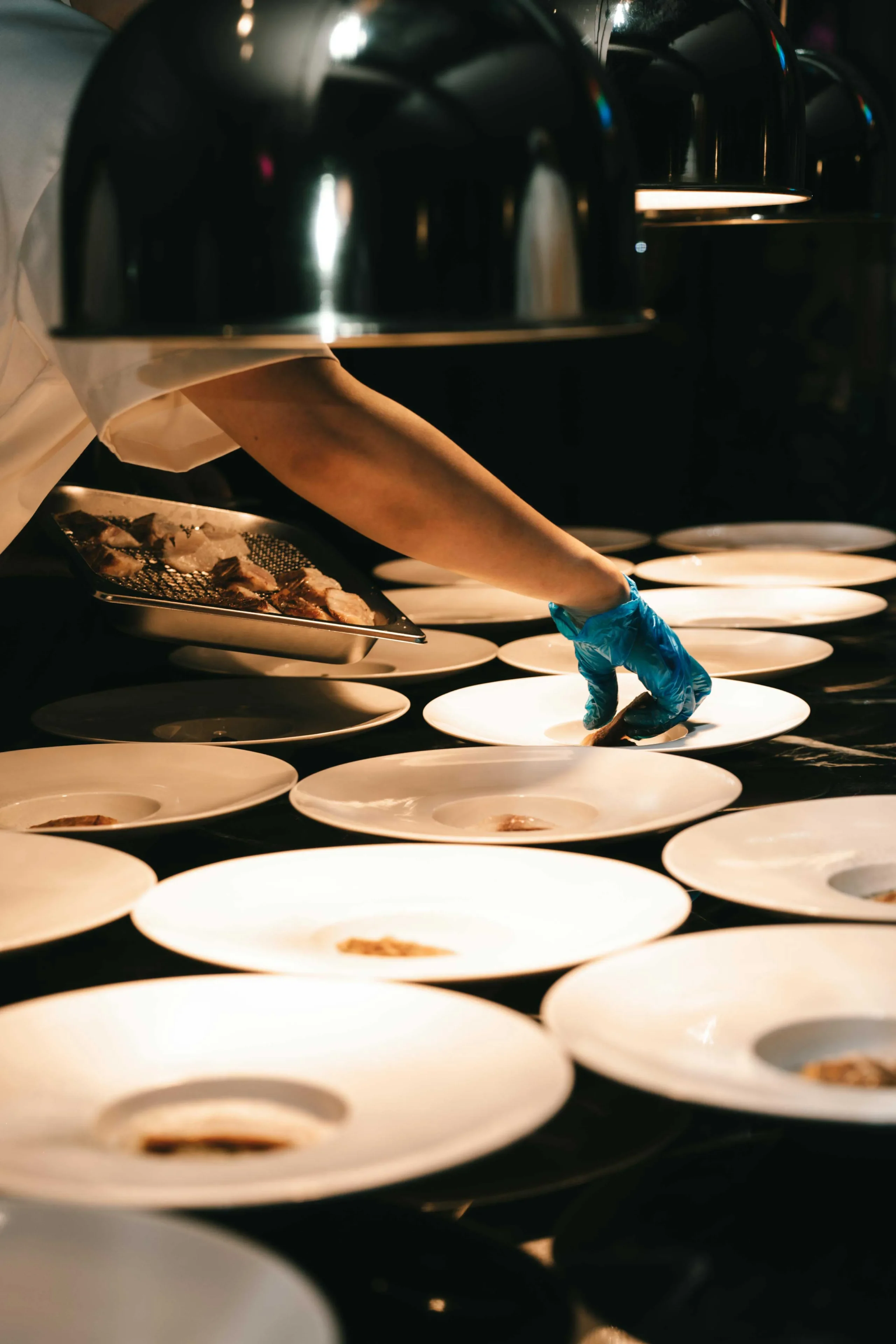 Chef wearing a blue glove plating small portions of food onto round white plates in a dim kitchen environment.