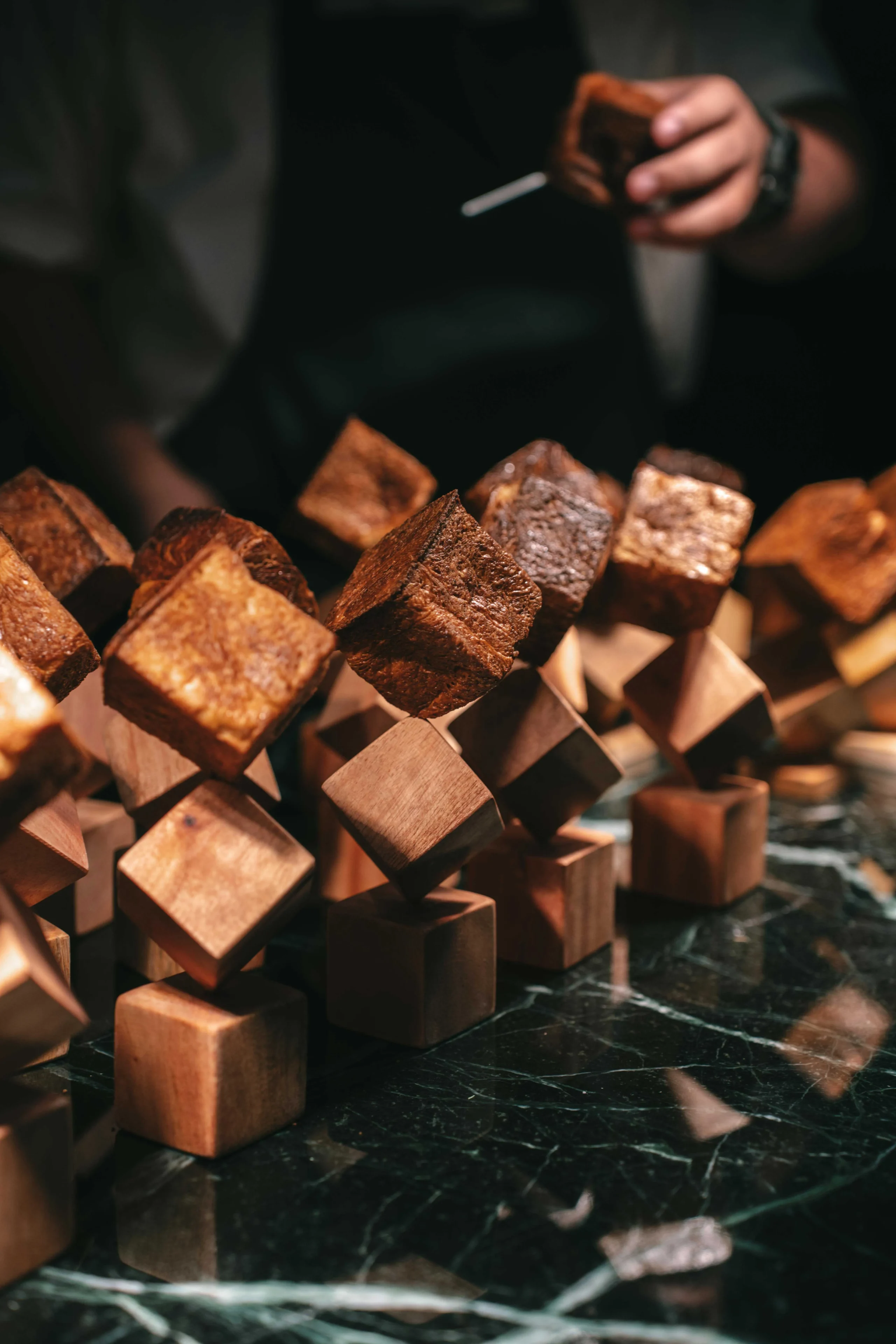 Close-up of brown, cubed chocolate treats threaded on wooden skewers in a geometric display on a dark, glossy surface. A blurred person holds a cigarette in the background.