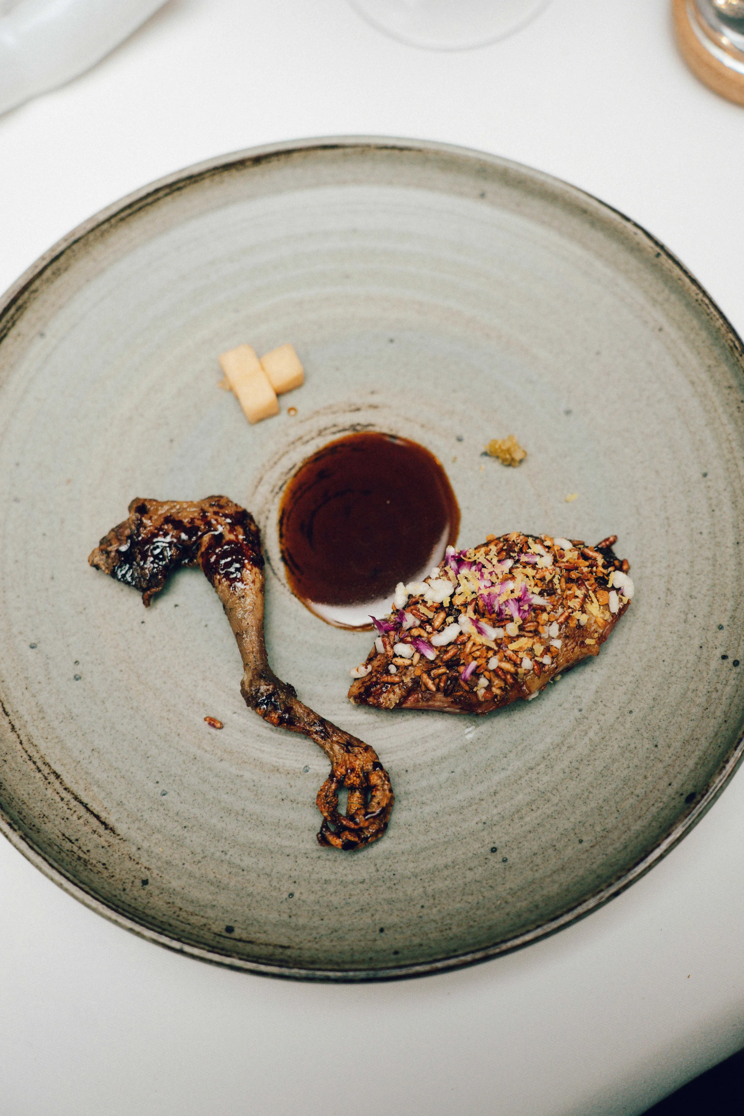 Grey ceramic plate holds a crusted, seed-coated meat piece, a small roasted chicken leg, and a pool of dark sauce in the center.