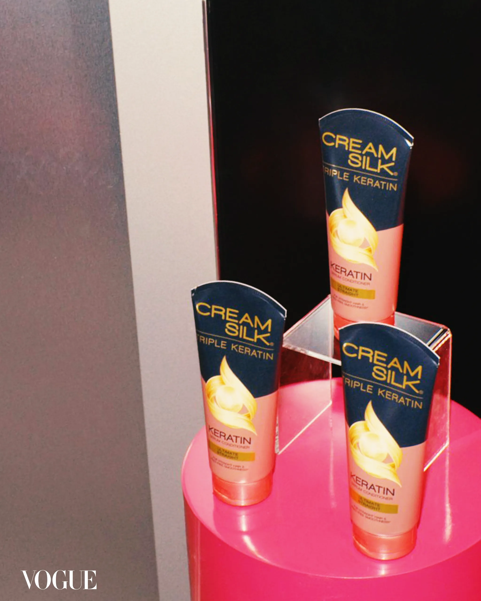 Pink circular display stand with multiple Cream Silk Triple Keratin hair care tubes on a black backdrop; Vogue watermark visible in the bottom-left corner.