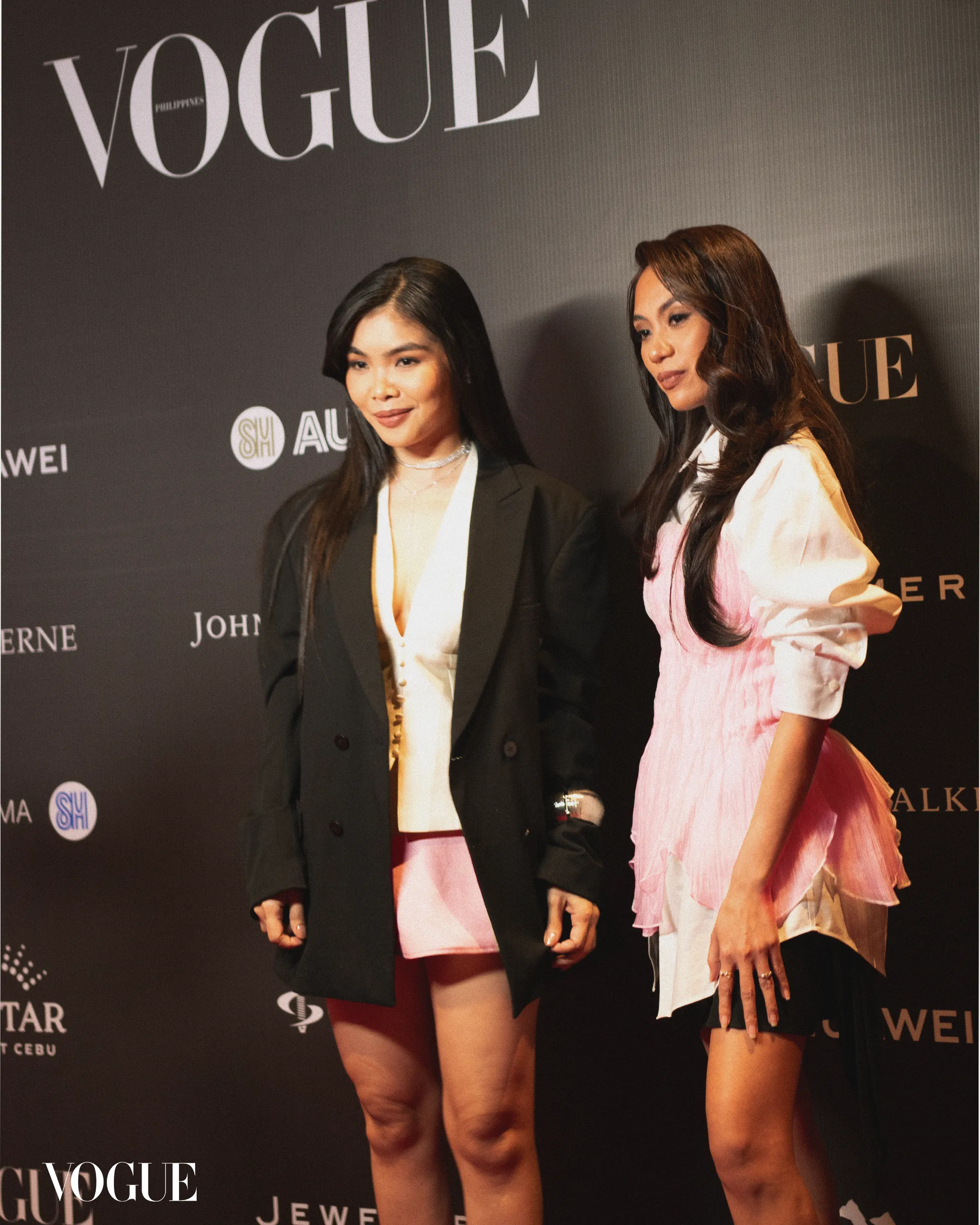 Two women pose together on a fashion event red carpet with a Vogue backdrop behind them.