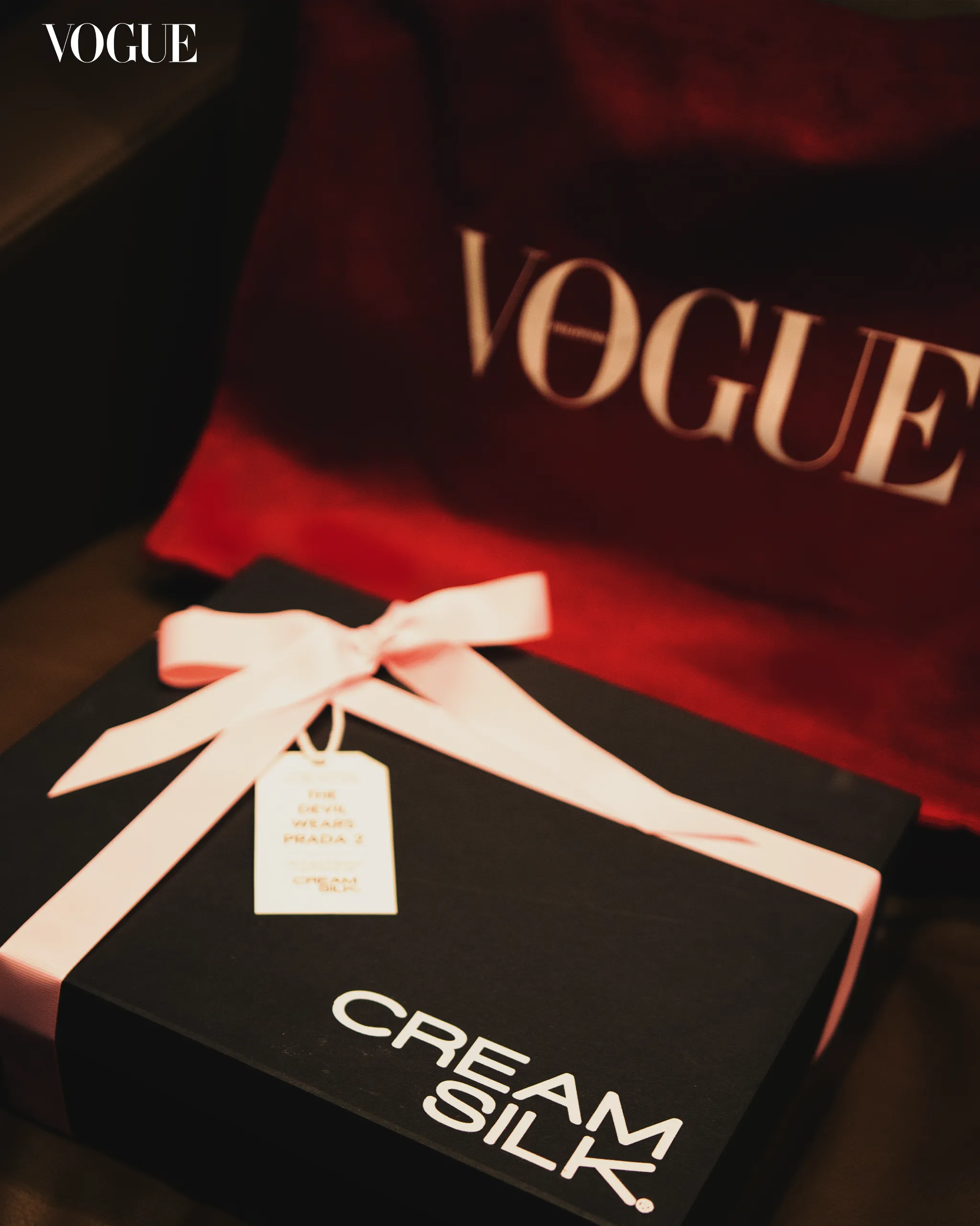 Gift box on a dark surface with a pale ribbon and tag, set against a red backdrop with a Vogue logo in the background.