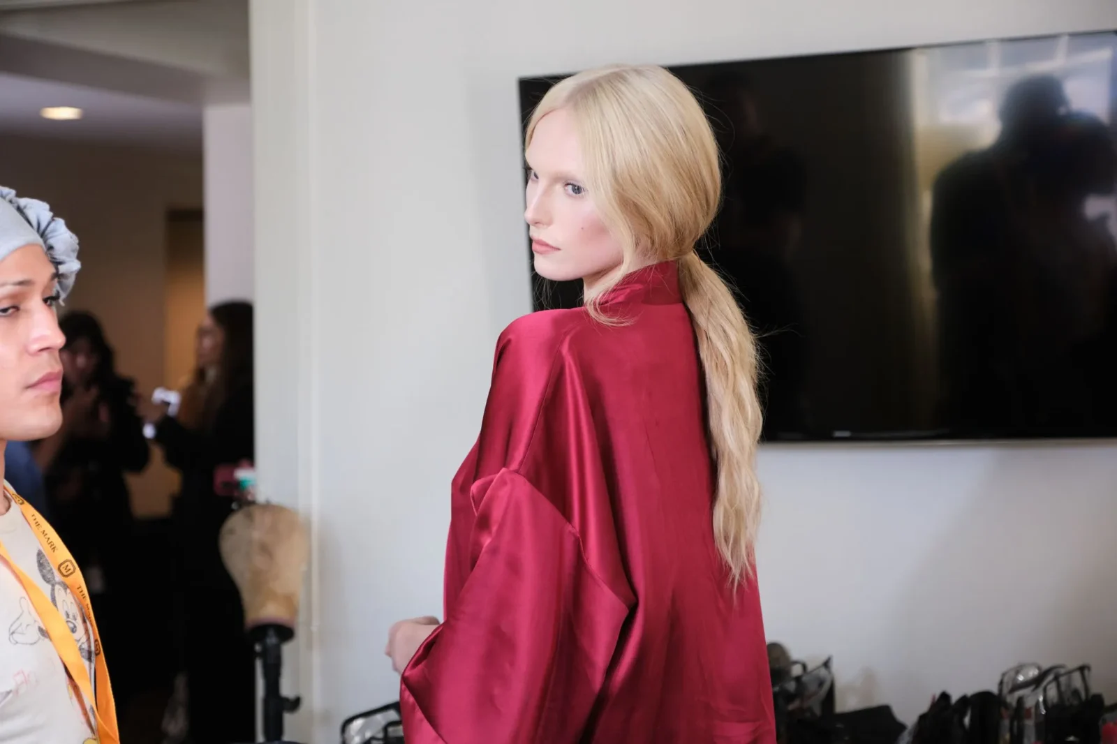Side view of a blonde model in a red satin outfit backstage, with a large screen on the wall in the background.