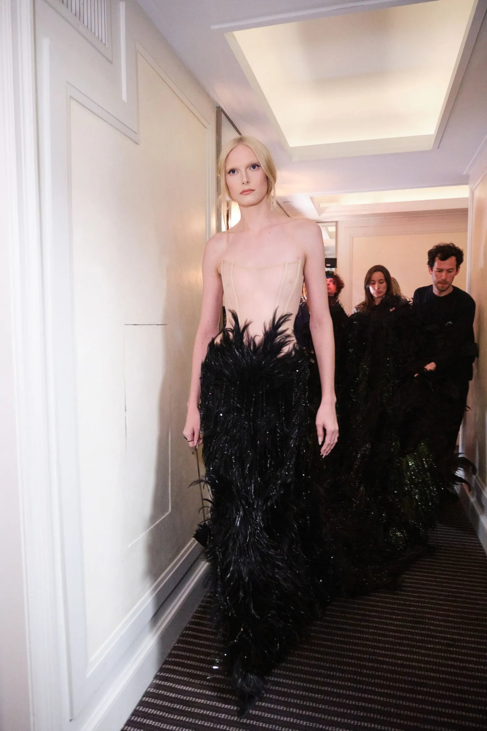 Model walking in a beige strapless corset dress with a black feathered gown in a hallway, backstage with crew in background