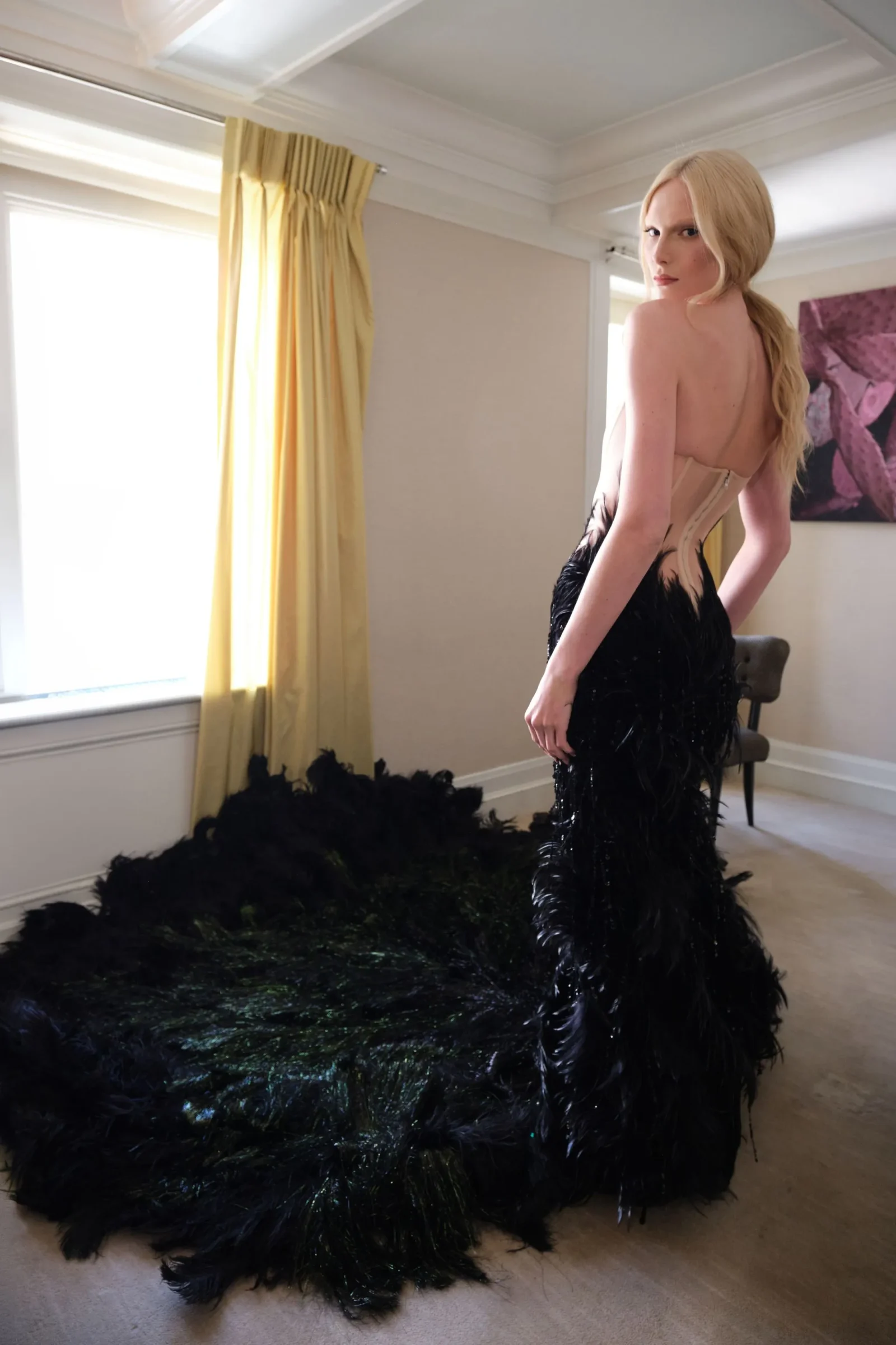 Blonde woman in a backless beige gown posing in a bright room with a large black feathered train on the floor.