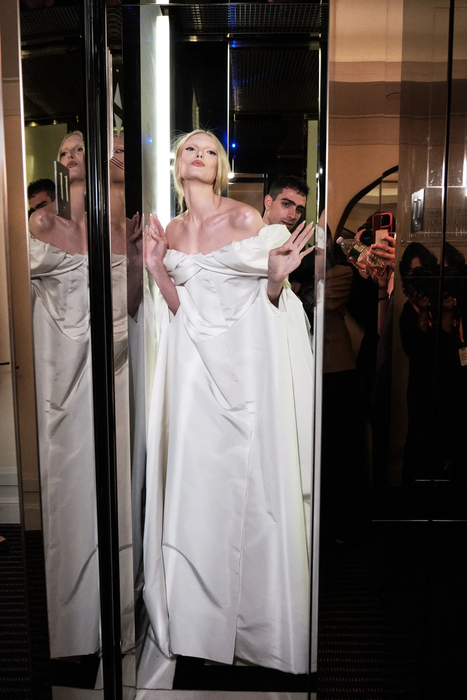 Woman in a white off-shoulder gown posing in an elevator, with a man behind her holding a water bottle and a reflective surface around them.