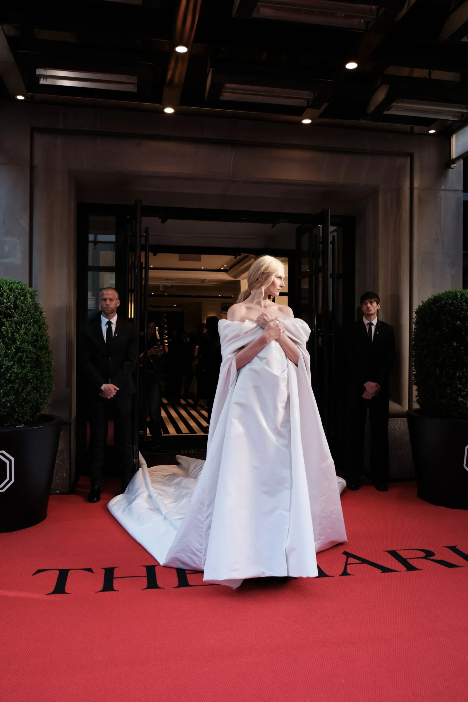Model in a white, voluminous gown with a long train posing on a red carpet at a venue entrance, flanked by suited security guards.