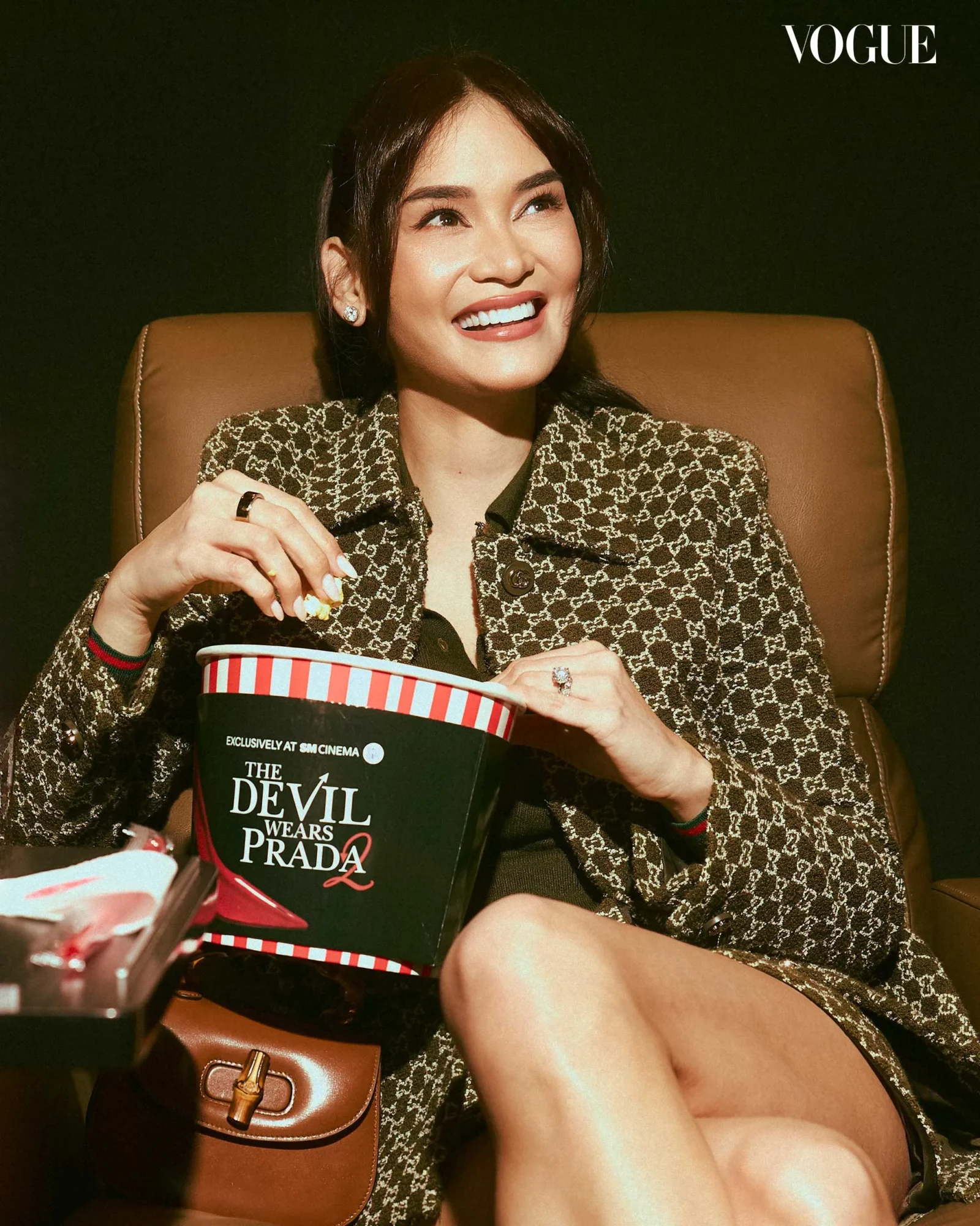 Smiling woman in a patterned coat sits in a brown chair, holding a striped popcorn bucket with The Devil Wears Prada logo; Vogue logo in corner.