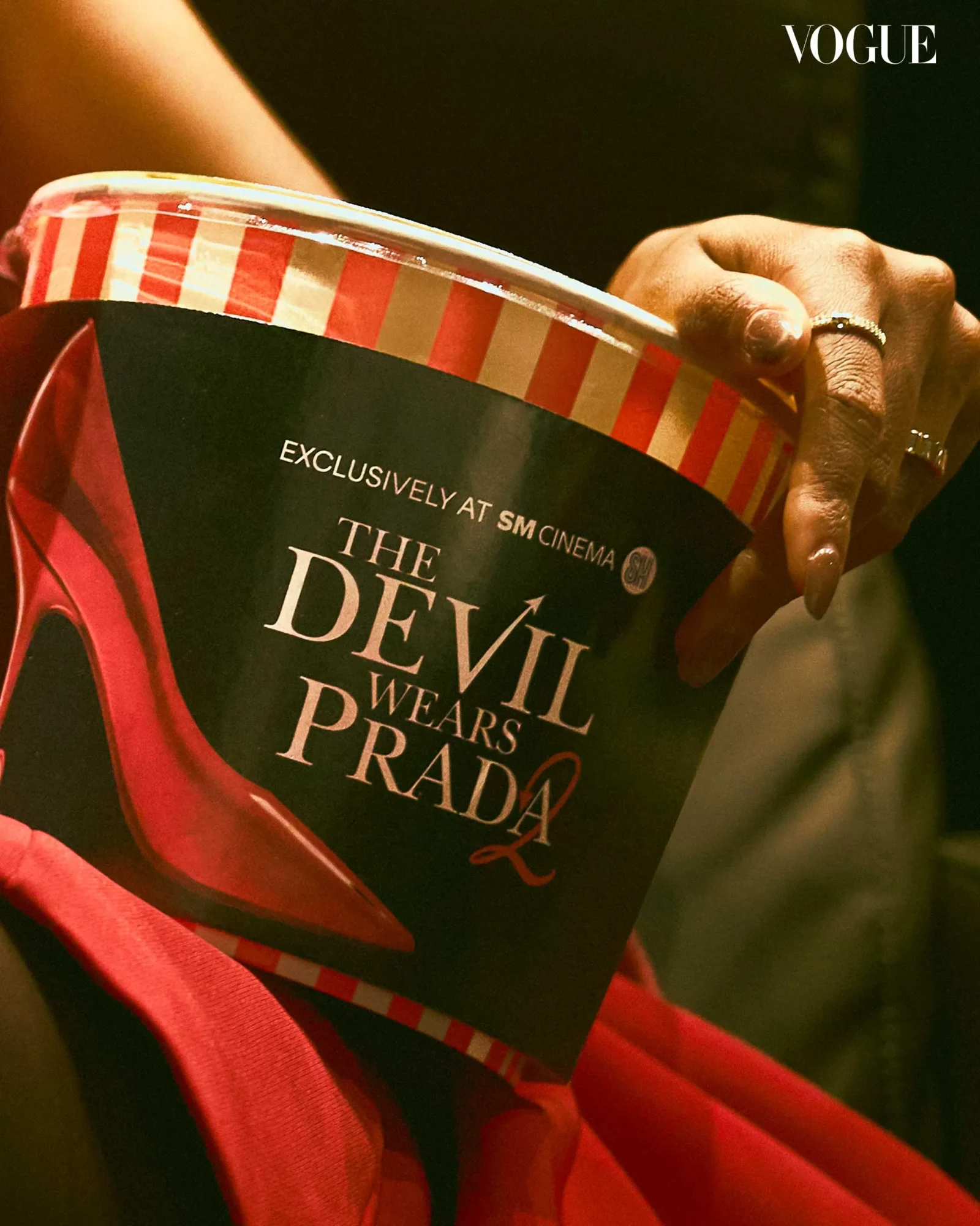 Close-up of a hand with rings gripping a black copy of The Devil Wears Prada, with a red striped border and red fabric nearby.