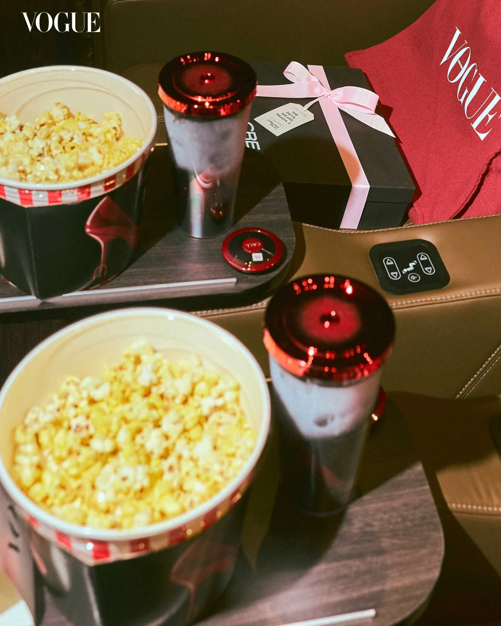 Vogue-branded scene with popcorn buckets, a red-lidded tumbler, and a black gift box tied with a pink ribbon atop a car console.