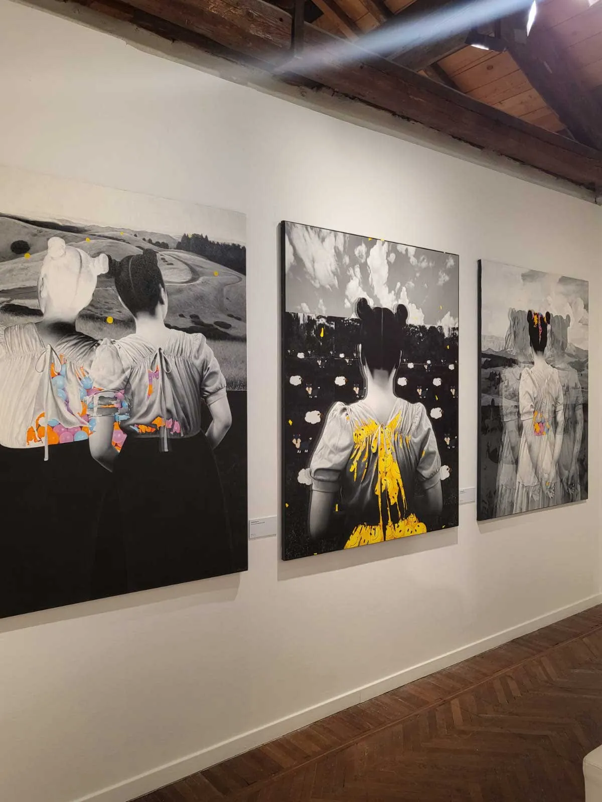 Three large black-and-white portraits of women viewed from behind, with colorful paint accents on their clothing, hanging in a gallery.