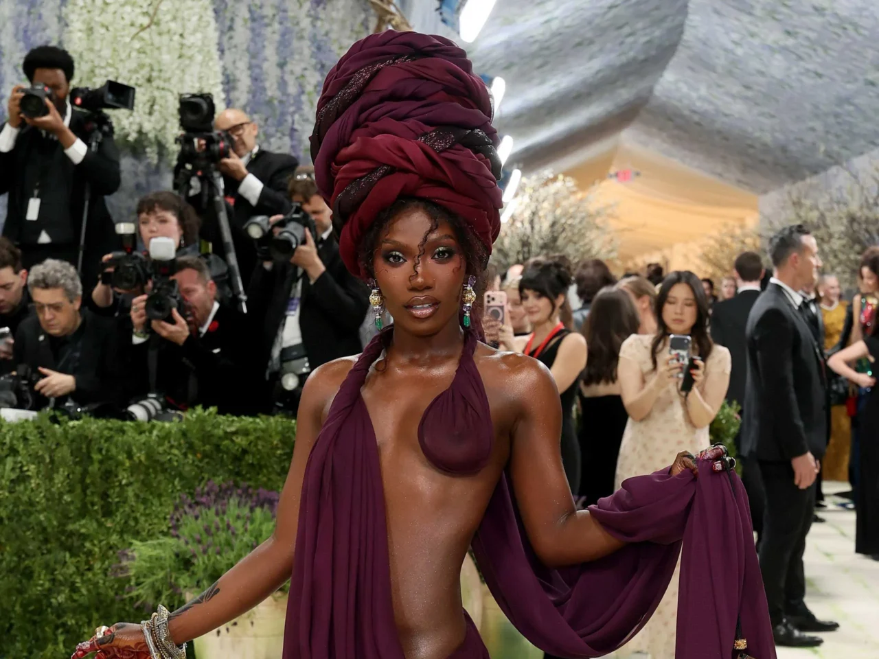 Woman wearing a burgundy turban-style headwrap at a red-carpet event, surrounded by photographers taking pictures.