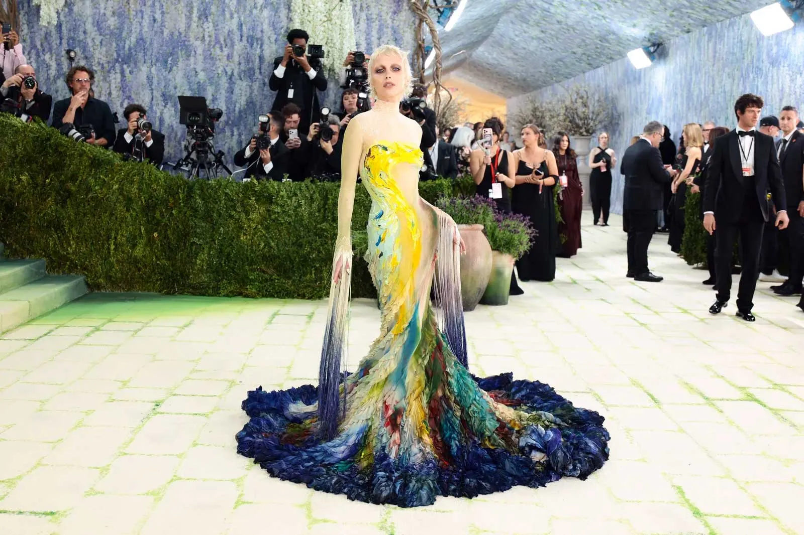 Model on a green-tiled runway wearing a bright, multicolored gown with a long, flowing train while photographers surround the scene.