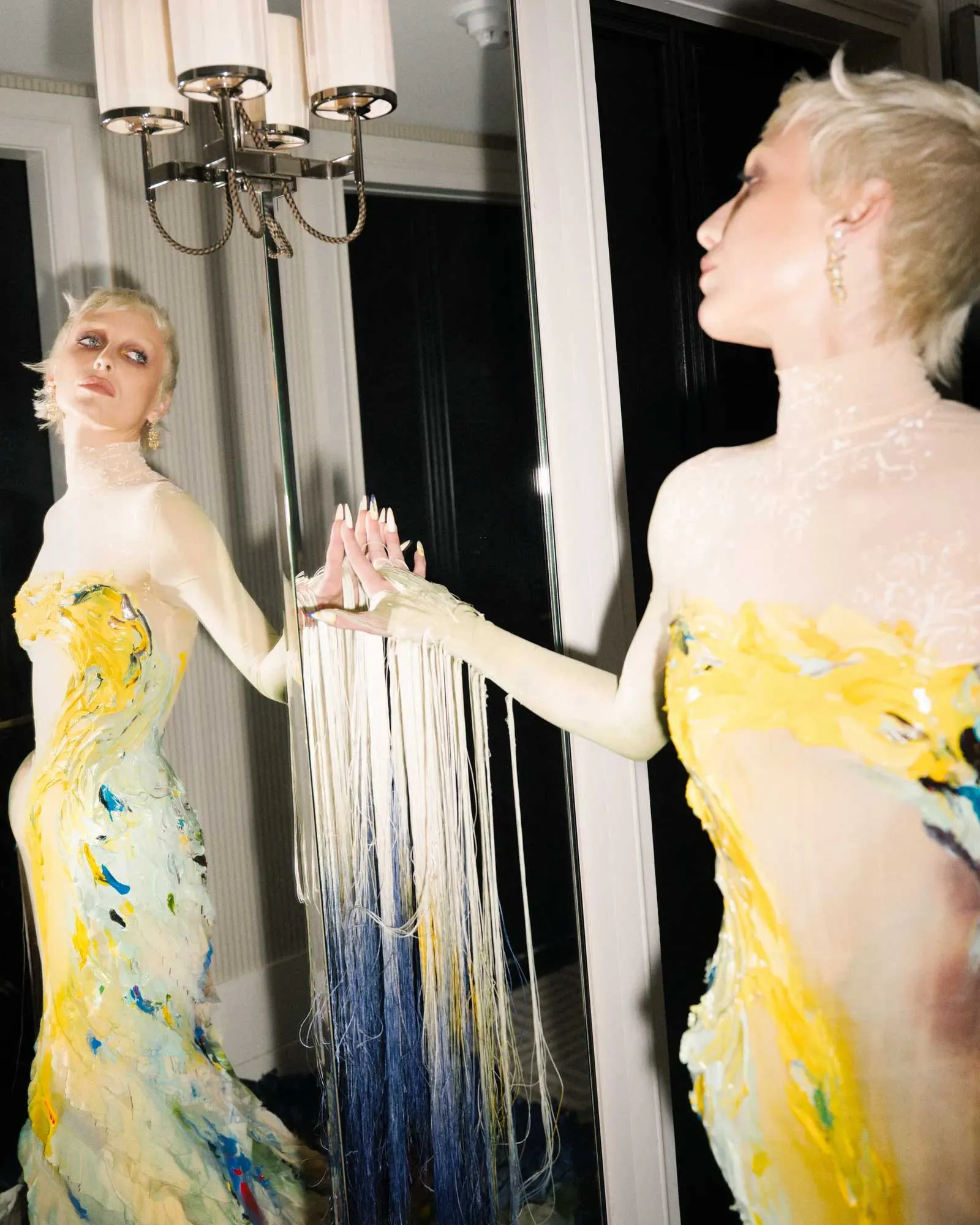 Person with short blond hair in a pale, paint-splattered gown stands by a mirrored wall, looking at their reflection under a chandelier.