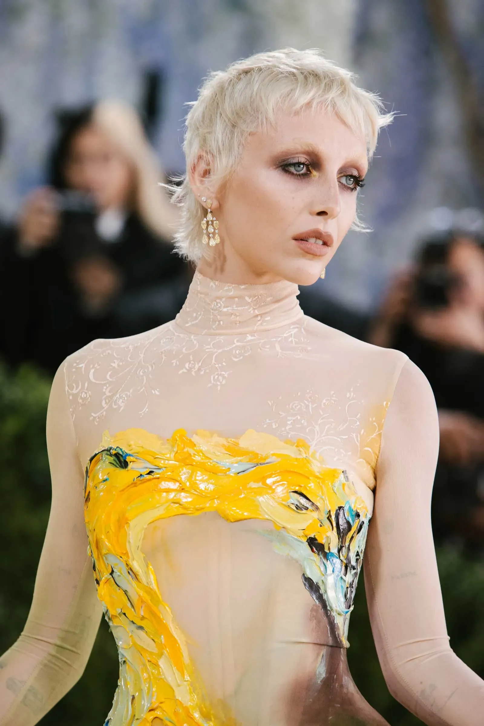 Person with short platinum blonde hair wearing a pale lace dress and dangling earrings, with bright yellow floral body paint across the chest and torso, outdoors.