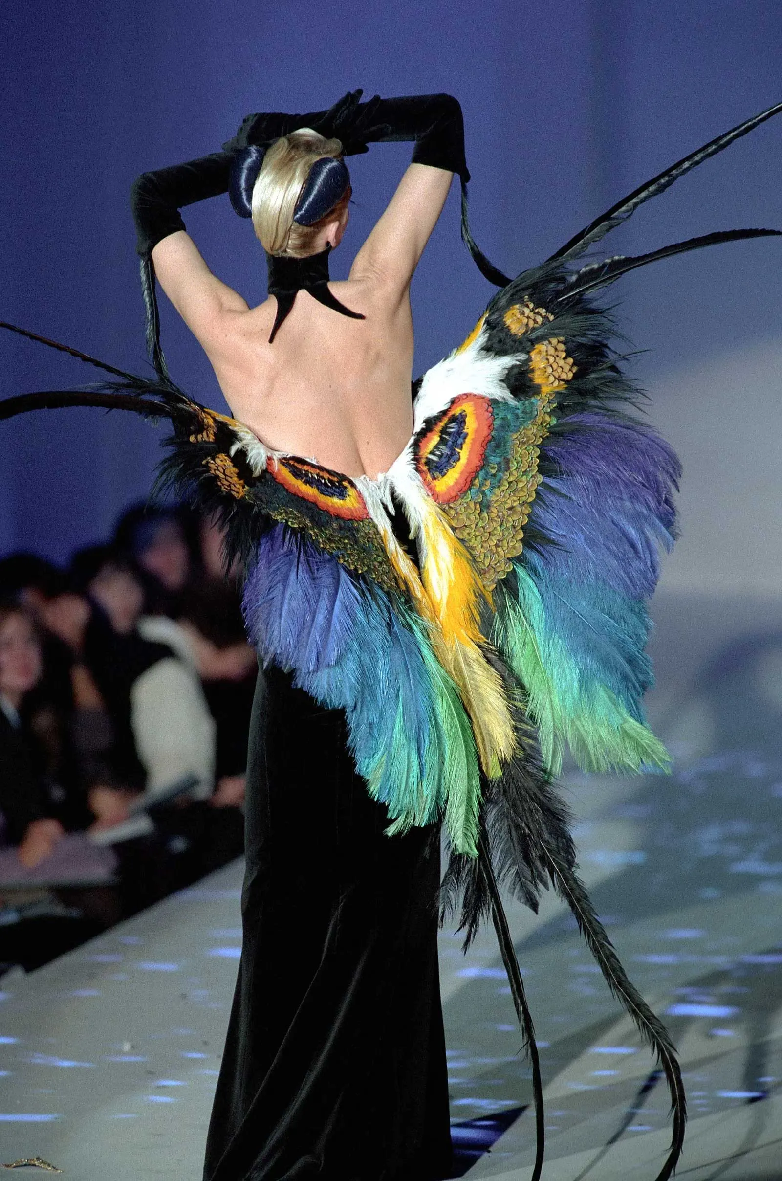 Back view of a fashion model wearing a dramatic multicolored feathered cape or wings attached to her waist during a runway show.