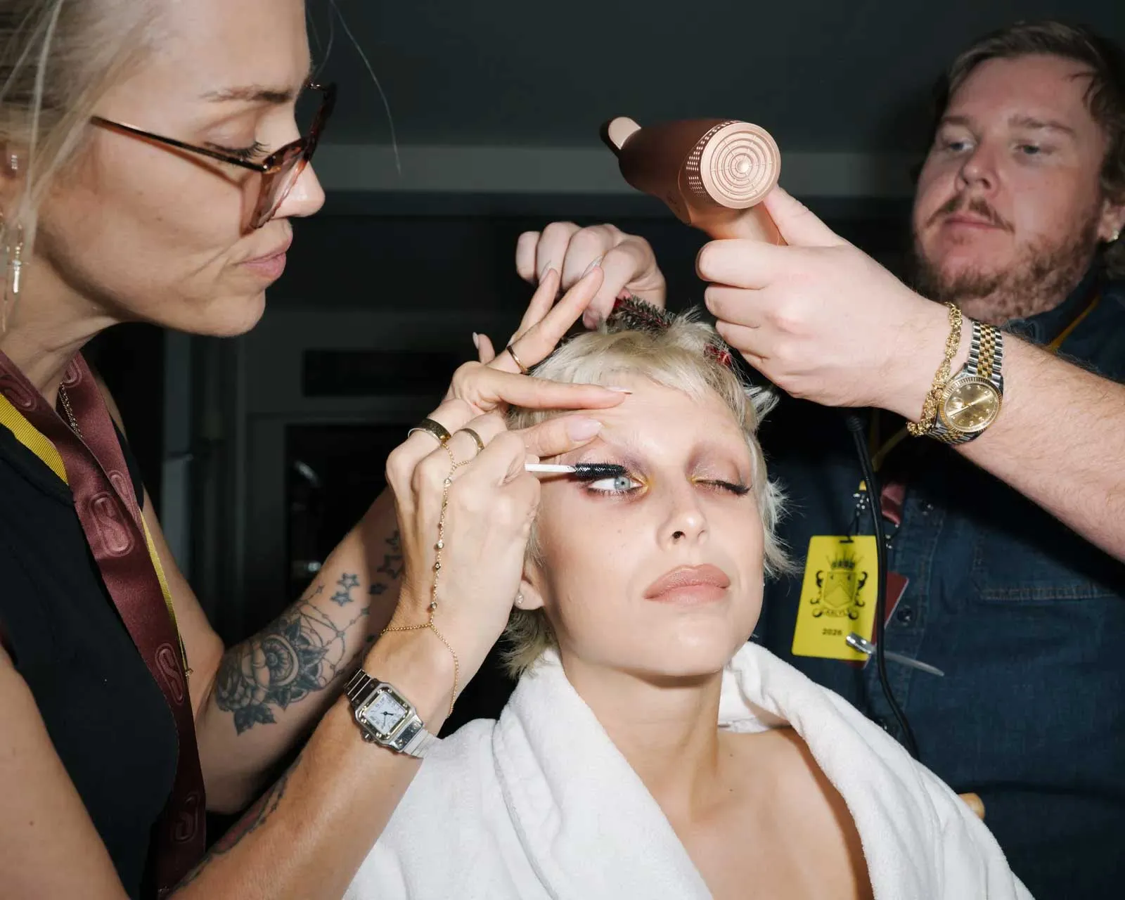 Makeup artist applies dramatic eyeliner to a seated model; others style hair with a curling iron nearby in a backstage setting.