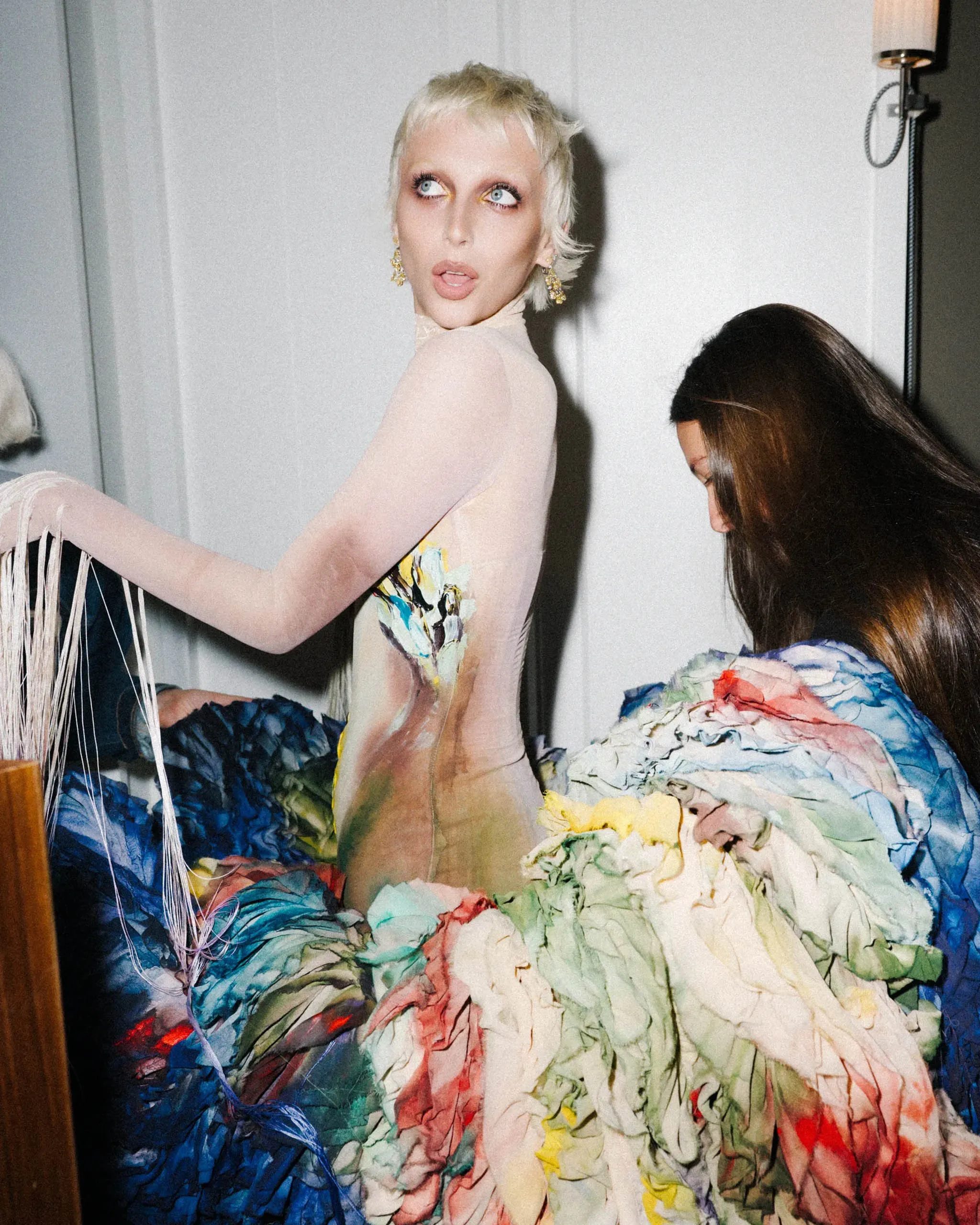 Model in a pale, sheer bodysuit with a large, multicolored crinkled fabric skirt, standing in a room as another person arranges the fabric nearby.