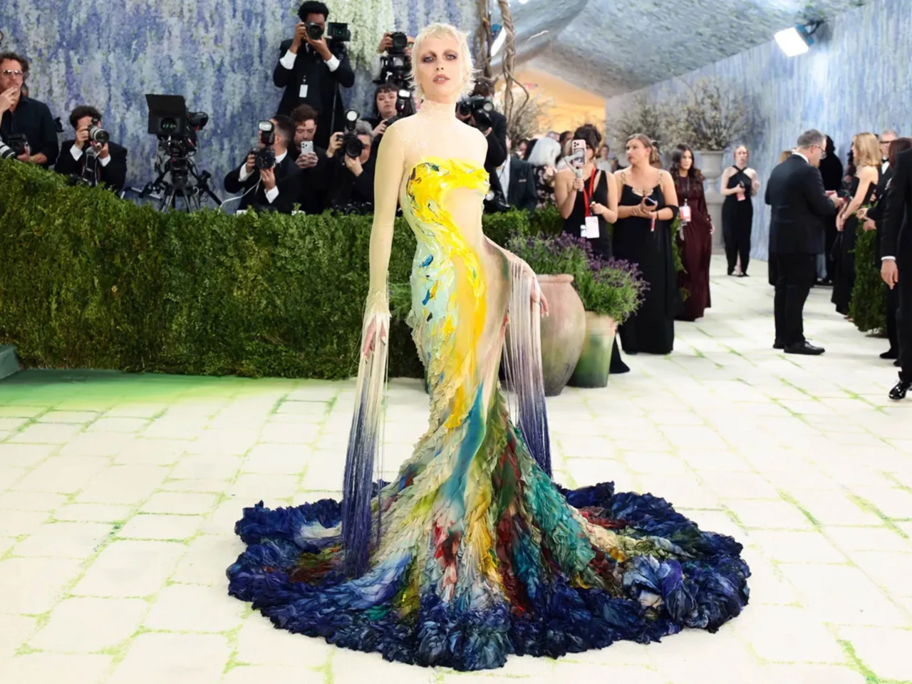 Model in a multicolored, flowing gown poses on a light-green tiled runway as paparazzi snap photos in a glamorous venue.