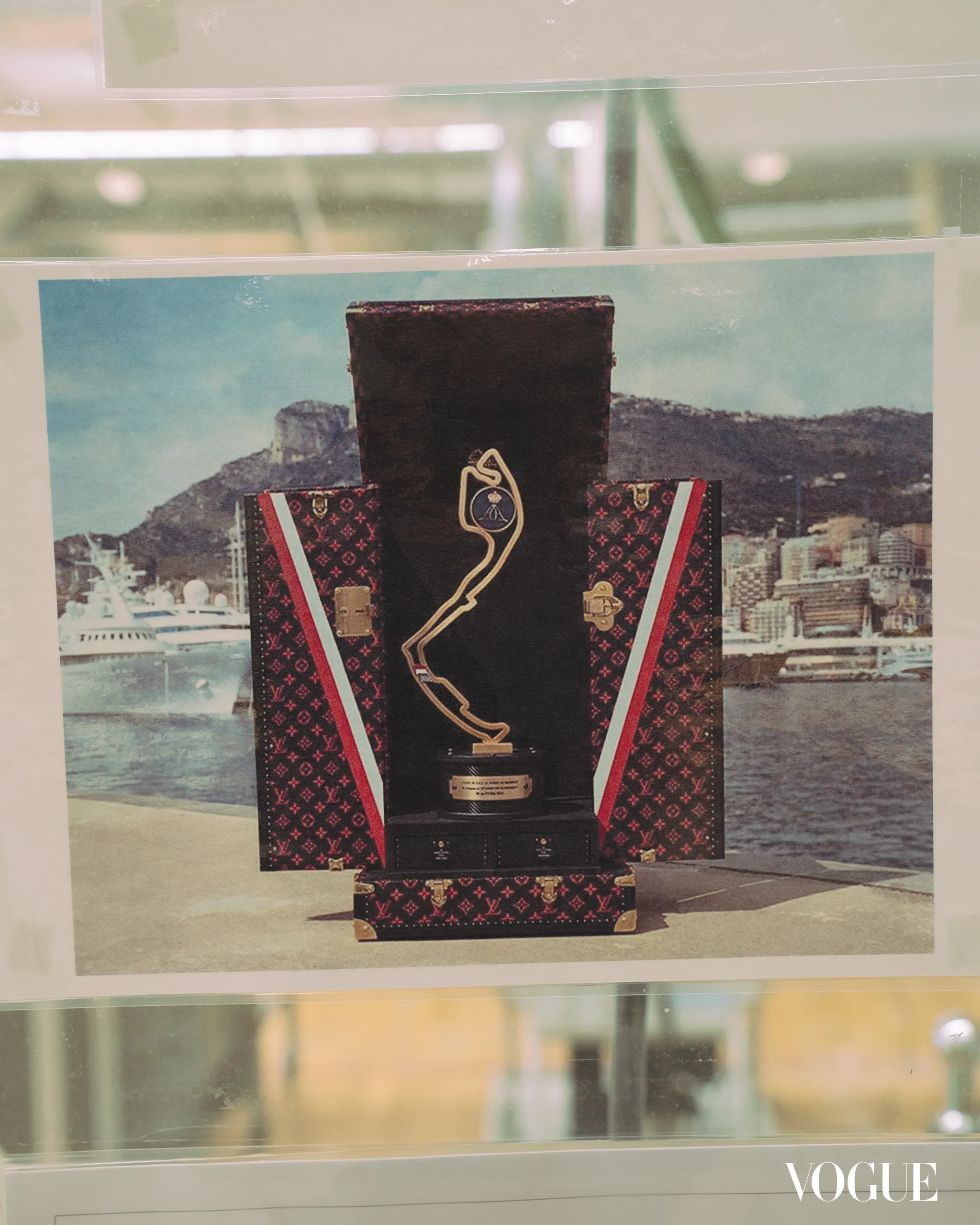 Vogue advertisement showing a tall black sculpture wrapped in Louis Vuitton luggage patterns at a harbor backdrop.